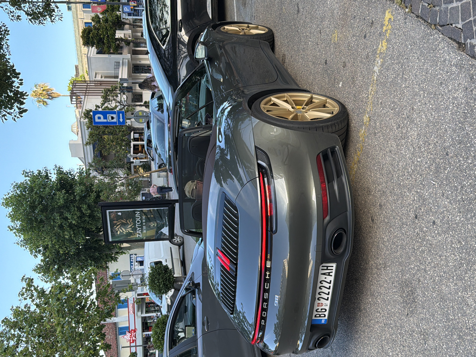 The image presents a daytime street-level view of a parking area on Mediteranska Street in Budva, Montenegro. In the immediate foreground, facing away from the viewer, is a dark metallic grey-green Porsche 911 convertible (992 generation) featuring prominent gold multi-spoke alloy wheels. The car displays a Serbian license plate, "BG 2222-AH," with "SRB" and "БГ" (Cyrillic for BG) text. The rear of the Porsche includes a full-width LED taillight strip and an engine cover with horizontal slats incorporating two vertical red accents. Two circular exhaust pipes are visible at the bottom of the rear bumper. A man wearing a baseball cap is seated in the driver's seat.
Behind the Porsche, a dark grey sedan is parked. To the left, a portion of another grey sedan is visible. The ground is dark asphalt with faded yellow parking space markings and a cobblestone curb on the right.
In the midground, several elements are discernible:
* A large, vertical black-framed billboard advertising "ZAYTOUN - THE TASTE OF THE EAST," with white text and a green olive branch graphic.
* A blue rectangular parking sign with a white "P," a white symbol of a hand holding a coin, and a left-pointing arrow, indicating paid parking in that direction.
* A woman in a light-colored top and dark pants is visible walking near the Zaytoun billboard.
* A white commercial van, resembling a Mercedes Sprinter, is parked further in the background.
* Various green trees, including large deciduous trees and smaller ornamental shrubs, are interspersed throughout the scene.
In the background, architectural structures dominate:
* A building with a reddish tiled roof, a light-colored facade, and distinctive blue window shutters. A red star logo with "Plazma" text is prominently displayed on this building, indicative of a store.
* Behind this, a more contemporary, multi-story building with a light-toned facade, balconies, and vertical dark slats. A distinct palm tree is growing on the rooftop of this building.
* Further to the right, a red and yellow flag, consistent with the flag of Montenegro, is visible on a pole.
* The sky above is clear and light blue, indicating fair weather conditions.