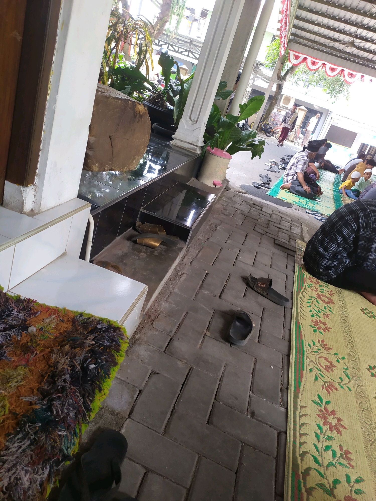 The image captures an outdoor scene in Sleman, Indonesia, likely a mosque courtyard or public prayer area, with several people engaged in prayer or rest on prayer mats. In the foreground, a colorful, shaggy rug is visible on a white tiled step. To the right, a yellow prayer mat with a floral pattern is laid out on a brick-paved ground. Scattered on the ground are several pairs of sandals, including a dark flip-flop and a black slide sandal.
Further back, more people are seated or kneeling on prayer mats, some wearing white caps and modest attire. In the background, there are architectural elements like white pillars and a red and white patterned awning. Green plants are present in pots, and a large log sits near a tiled area, suggesting a blend of natural and constructed elements. The lighting indicates it is daytime, with sunlight filtering through the structures and foliage. The overall atmosphere appears to be one of communal gathering and religious observance.