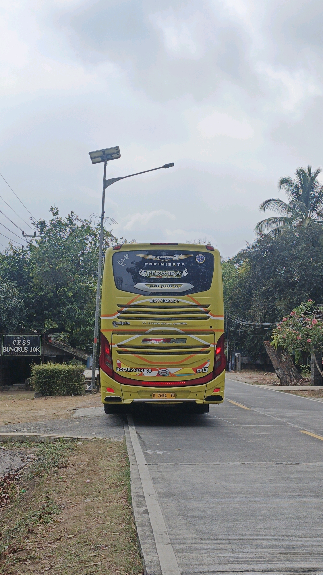The image shows the rear of a yellow tour bus parked on the side of a road in Cibenda, Indonesia. The bus has "PARIWISATA PERWIRA" and "adi pulro" written on its rear window. There is a sign on the left side of the image that reads "CESS BUNGKUS JOK". A street light with a solar panel is visible in the background. The environment appears to be rural or suburban, with trees and foliage surrounding the road. The sky is cloudy.
