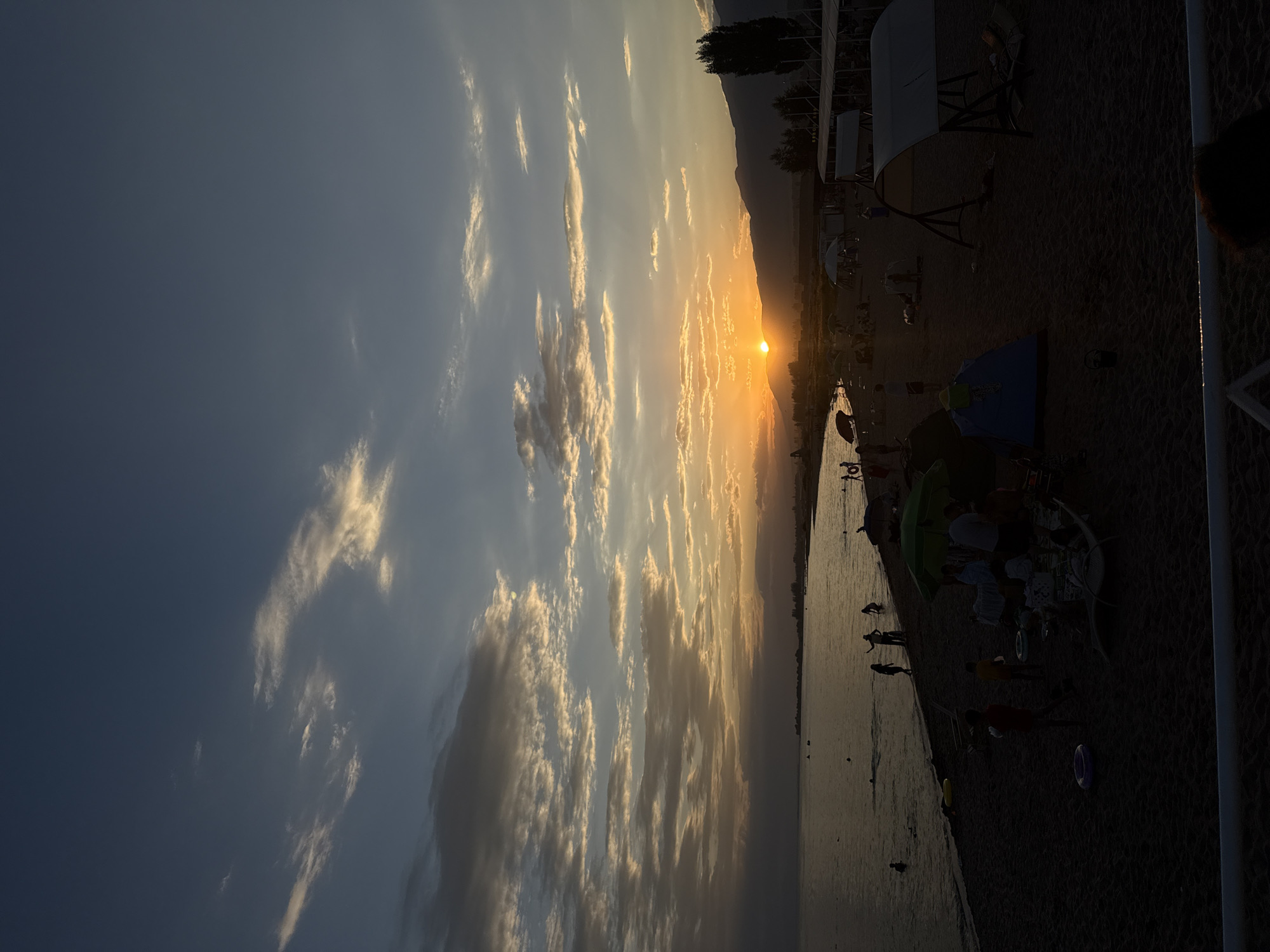 This is a full shot of a beach scene in Sary-Oy, Kyrgyzstan, during sunset. The sky is a gradient of deep blue at the top, transitioning to lighter blues and then to warm oranges and yellows near the horizon, where the sun is setting behind a mountain range. Scattered clouds, illuminated by the setting sun, add texture to the sky.
The foreground is a dark sandy beach. To the left, a body of water is visible, with a few silhouetted figures swimming or standing in the shallows. The water reflects the colors of the sky. Further into the scene, the beach is populated with various elements. There are several umbrellas providing shade, one of which is bright green. Tents, including a blue one, are set up on the sand. Beach chairs are scattered around, some occupied. Silhouetted figures of people are engaged in activities such as walking, sitting, and interacting. Some appear to be children playing. In the distance, along the coastline, there are some structures and trees. The overall impression is a lively, end-of-day scene at a popular beach resort.