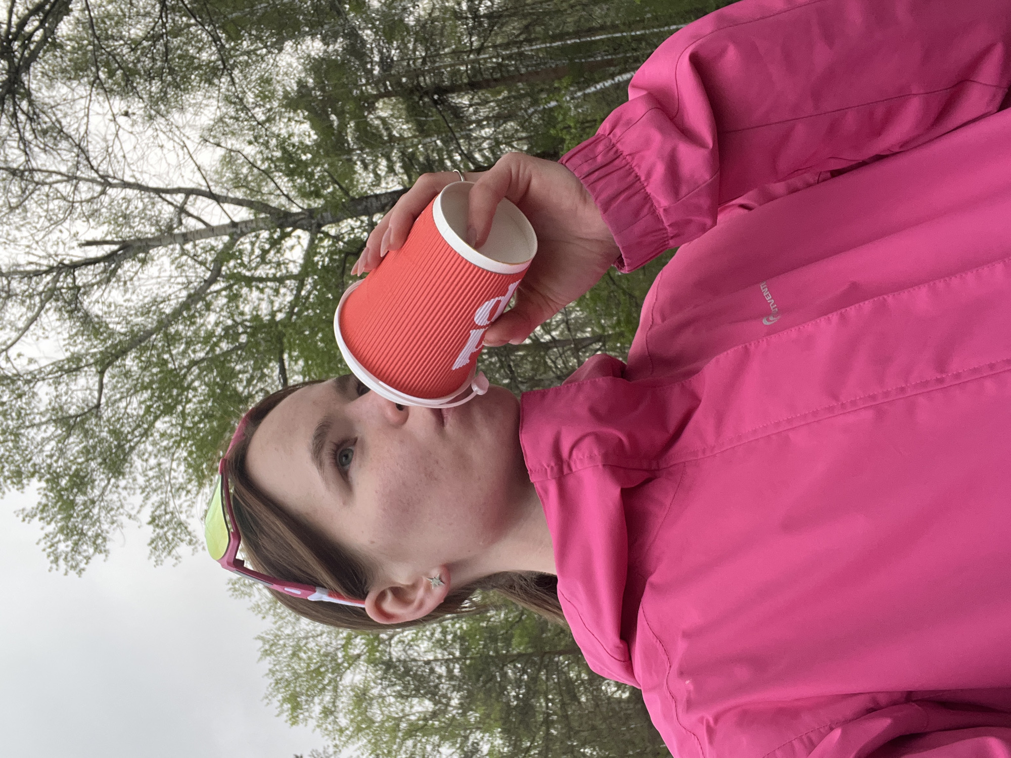 A young adult female in a pink windbreaker is seen in an outdoor environment, possibly a park or wooded area. She is drinking from a red paper cup, which she holds in her right hand. She is wearing sunglasses atop her head and a silver star-shaped earring in her left ear. The background shows bare trees and a cloudy sky. The location cannot be determined from the image.