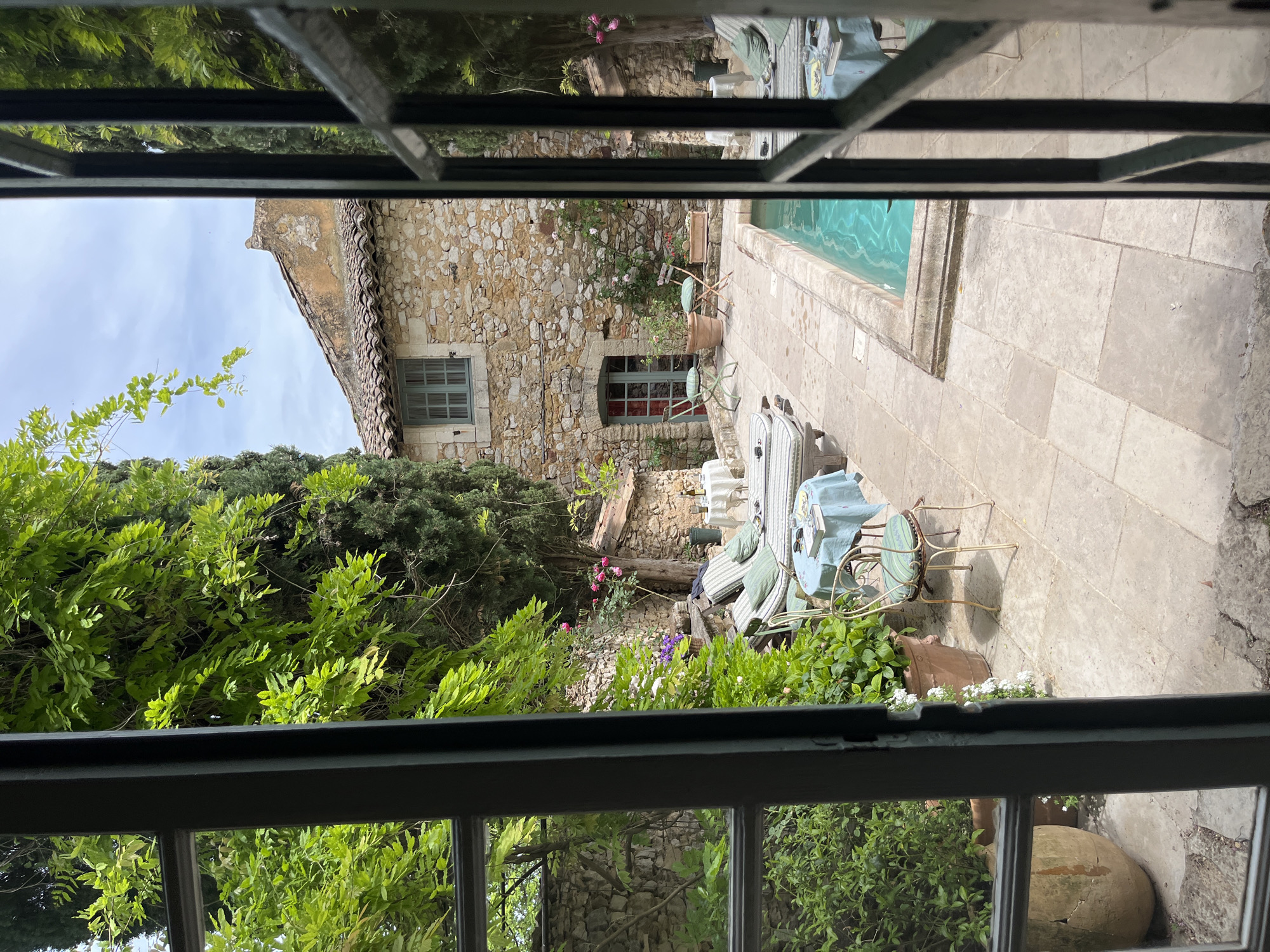 The image presents a tranquil outdoor scene, viewed through the panes of a window, looking out onto a charming courtyard.
In the midground, a traditional stone house with a tiled roof stands, showcasing rustic architecture with light-colored, irregular stones. It features a window with closed green shutters on the left side and a smaller window below it, revealing a darker interior.
To the right of the house, a rectangular swimming pool with inviting turquoise water is set into a paved patio. The patio is made of light-colored flagstones, extending across the foreground and along the pool's edge.
The courtyard is rich with lush greenery, including tall cypress trees, leafy vines, bushes, and flowering plants, some with small pink blossoms. Several terracotta pots containing plants are scattered across the patio, enhancing the garden's appeal.
Outdoor furniture is arranged on the patio, suggesting a comfortable area for relaxation and dining. This includes two round tables: one draped with a light blue tablecloth, accompanied by several chairs with light-colored, possibly striped, cushions; and another smaller table with a striped top and two chairs, one with a green cushion. An additional armchair or lounger with a striped cushion is also visible.
The scene is illuminated by bright daylight, casting shadows from the furniture, indicating a clear and sunny day, likely late morning or afternoon. The sky visible above the house is partly cloudy. No people are present in the image, and no text is visible. The overall aesthetic of the stone architecture, Mediterranean-style vegetation, and sunny ambiance aligns with the specified location of Saint-Victor-la-Coste, France, evoking a sense of peaceful, leisurely outdoor living.