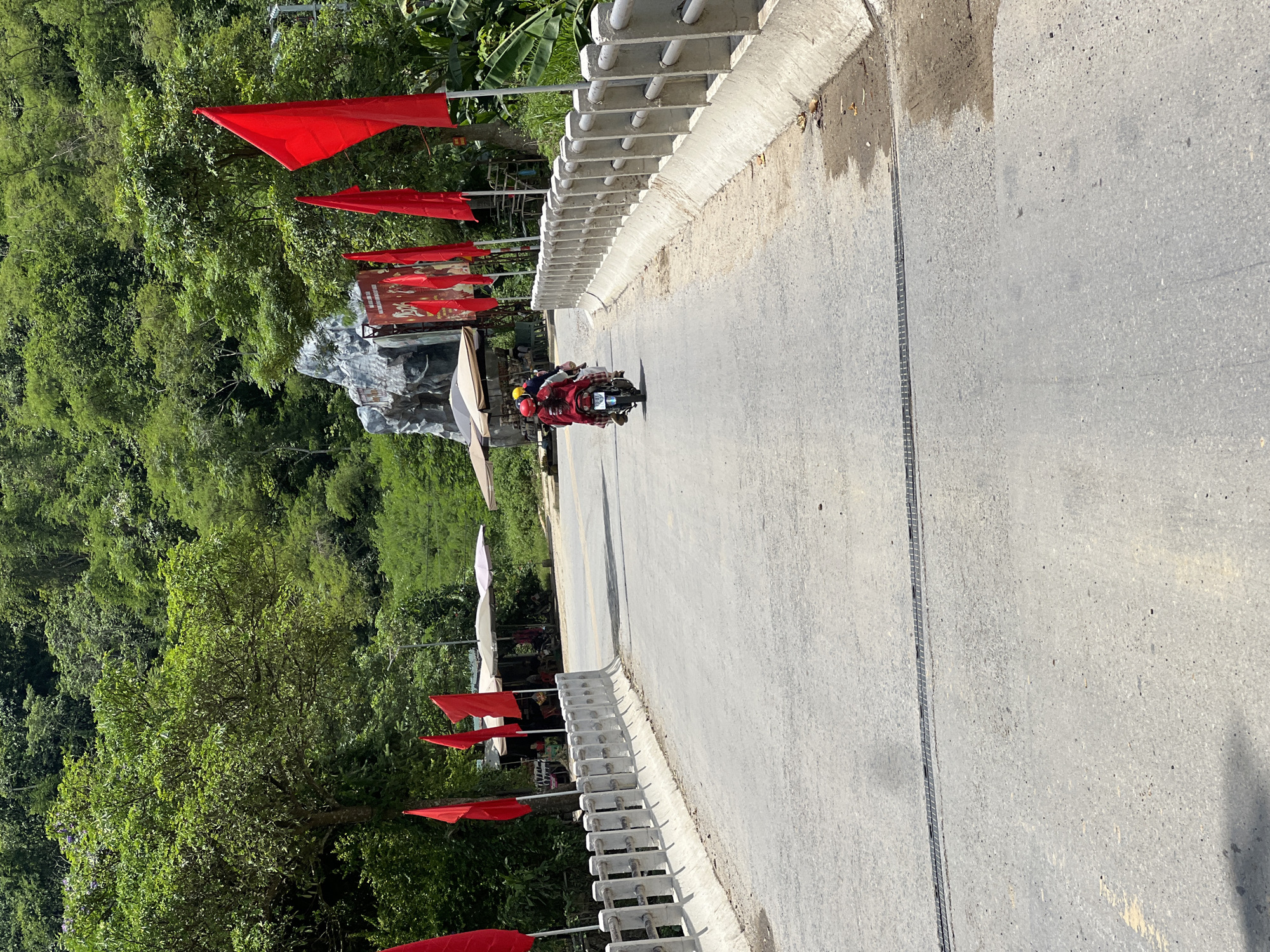In Minh Tân Commune, Vietnam, a road on a bridge leads towards a lush, green hillside covered in dense trees. Red flags flutter along the right side of the bridge, and a stone sculpture of a face is visible on the hillside. Two people on a red scooter, both wearing helmets, are traveling away from the viewer on the road. The bridge has a concrete barrier with horizontal railings on both sides. Various stalls and umbrellas are set up along the side of the road at the base of the hill, with some parked motorcycles visible. The road surface shows some wear and tear, with a metal grate running across its width.
