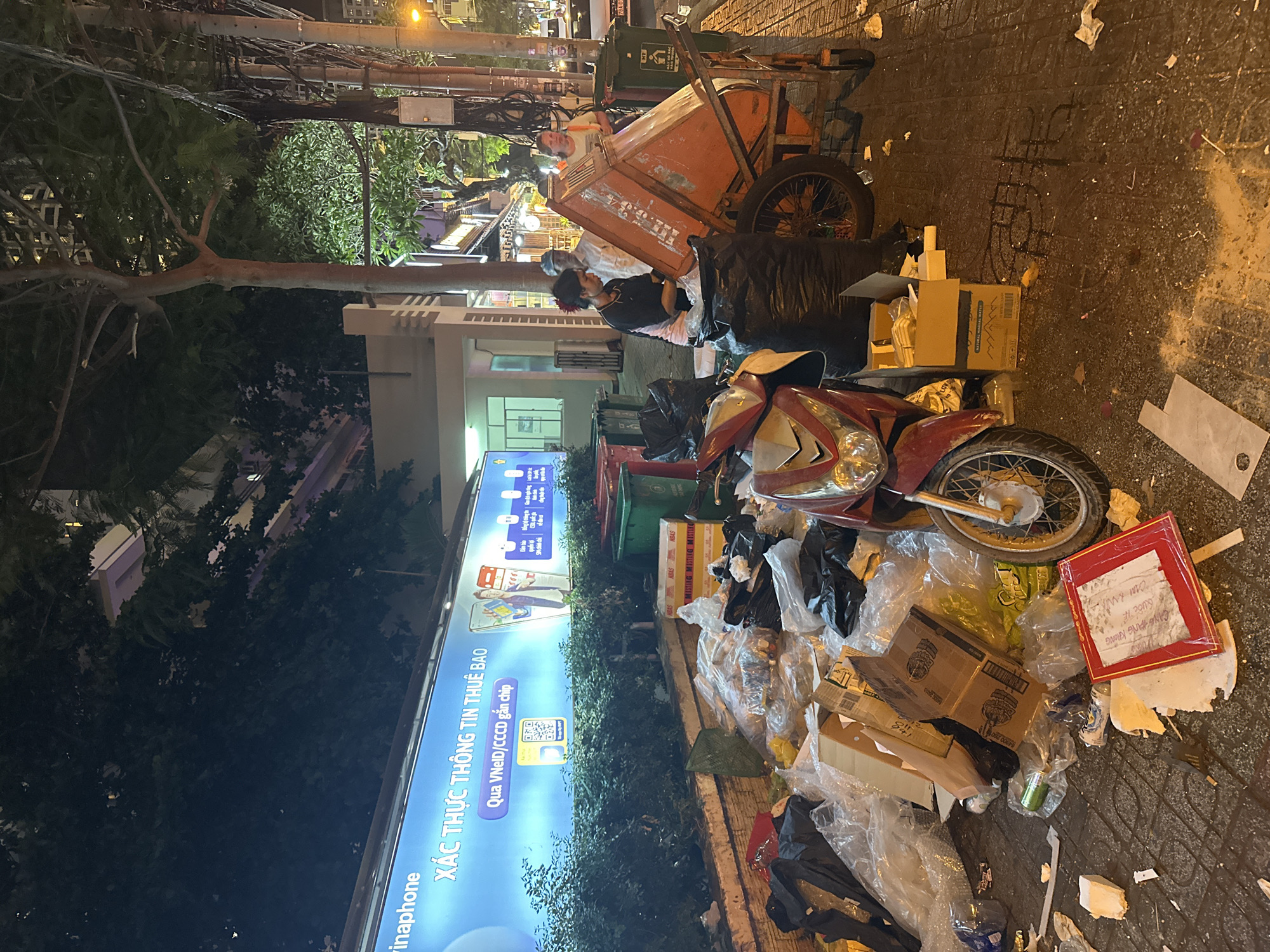 The image depicts a cluttered urban scene at night in Nha Trang Ward, Vietnam. A red and silver motorcycle is parked amidst a large pile of garbage bags, cardboard boxes, and loose trash on a paved street. In the background, a few people are visible near an orange garbage cart with large wheels. One person, wearing a dark shirt, is interacting with the garbage cart, possibly sorting or collecting trash. Another person in a white shirt stands nearby.
The lighting suggests it is nighttime, with streetlights and illuminated signs casting a warm glow on the scene. A large billboard with Vietnamese text and graphics is prominently displayed on the left side of the image, featuring the Vinaphone logo and text that translates to "VERIFY SUBSCRIPTION INFORMATION via VNeID/Chip-mounted CCCD". Other signs and lights suggest this is a commercial or residential area.
The overall impression is one of urban life and waste management, with the discarded items and the presence of a garbage cart indicating the collection or disposal of refuse. The abundance of trash suggests an untidy environment. A red framed note with handwritten text in Vietnamese is visible among the debris. The scene feels busy and active, despite the late hour, likely due to the activity surrounding waste collection.