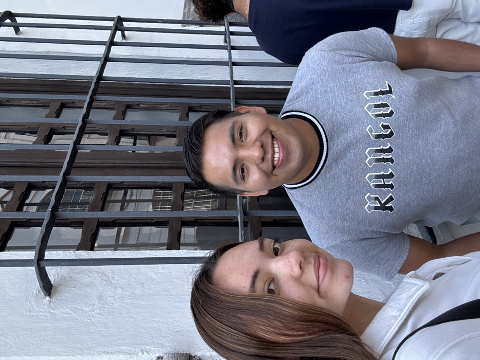 The image captures three individuals outdoors, positioned in front of a colonial-style building typical of the historic city center of Puebla, Mexico. On the left, a young female with long brown hair and fair skin wears a white collared shirt and has a black strap visible over her left shoulder. She gazes slightly to her right with a subtle smile. To her right, a young male with short dark hair and a medium complexion wears a grey t-shirt featuring a black and white striped collar and the word "KANGOL" in a stylized gothic font across the chest. He smiles directly at the camera, showing his teeth. On the far right, partially visible, is the back of a third individual with dark, curly hair, wearing a dark blue long-sleeved shirt and white pants or shorts. The background features a white, textured stucco wall with a prominent dark brown wooden window frame, segmented into multiple panes, and covered by a dark grey or black iron security grill. The scene is illuminated by natural daylight.