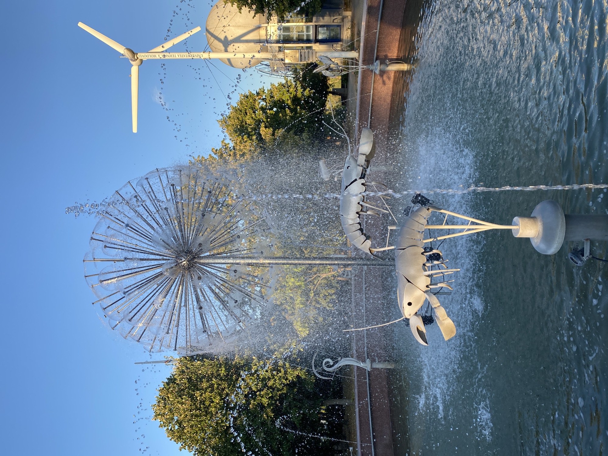 The image depicts an outdoor fountain with a modern design, featuring metallic sculptures of lobsters or crayfish as the focal point. These sculptures are positioned above the water, with one appearing to be closer to the viewer. The fountain itself is a large, spherical structure made of numerous thin metal rods radiating outwards, reminiscent of a dandelion seed head or a globe. Water is actively spraying from this central spherical element, creating a dynamic visual effect. In the background, several trees with green foliage are visible, suggesting a park or a green space. Further in the background, a white wind turbine stands tall against a clear blue sky. Adjacent to the wind turbine, there is a large, spherical building. The overall setting appears to be a public area, possibly a park or a university campus, given the presence of the wind turbine and the spherical building which might be a research facility or an observatory. The lighting suggests it is daytime, with the sun casting a warm glow on the scene. This scene is located at the Technical University of Moldova in Chișinău, Moldova, specifically in the courtyard area where the university has installed this unique fountain and educational equipment.