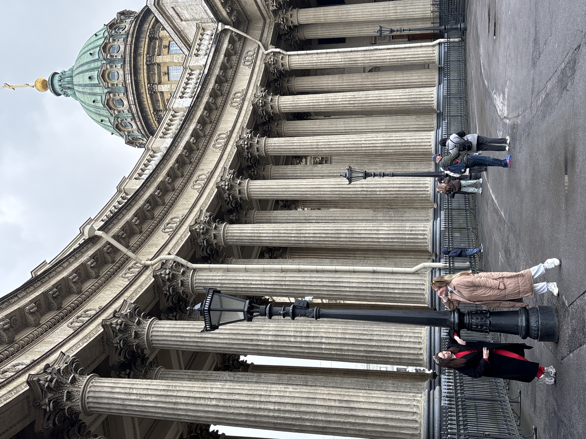 The image captures a low-angle perspective of the Kazan Cathedral in Saint Petersburg, Russia. The architecture features a large dome with a golden cross atop, supported by numerous classical columns. Several people are on the sidewalk along the building's base. One woman wears a long dark coat with a red garment underneath. Another woman wears a tan coat and light pants. Two people stand further down the sidewalk facing the building. One leans to look in their phone. A black, ornate street lamp stands between the people and the columns. The scene is outdoors on a day with overcast skies, and the pavement appears wet.