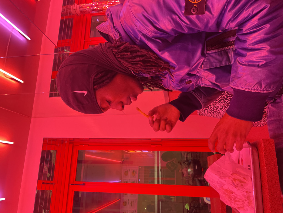 A person with dreadlocks wearing a black Nike hat and a puffy, blue jacket eats a french fry in a brightly lit, red-toned room. Reflected in a window is a scene of outdoor activity. A metal tray holding crumpled paper is positioned in front of the person. The setting appears to be inside a fast-food restaurant or similar establishment.
