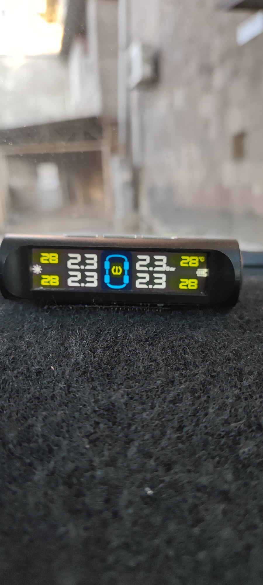 The image shows a tire pressure monitoring system (TPMS) display on a dark, fuzzy surface, possibly a car dashboard. The display reads "28" with a sun icon, "2.3", a tire symbol, "2.3", and "28°C" with a battery icon. All tires display 2.3 bar pressure. The background shows a blurred cityscape seen through a window. Based on visual similarities in architectural details, the image may have been taken in Yerevan, Armenia. There are no people visible in the image.