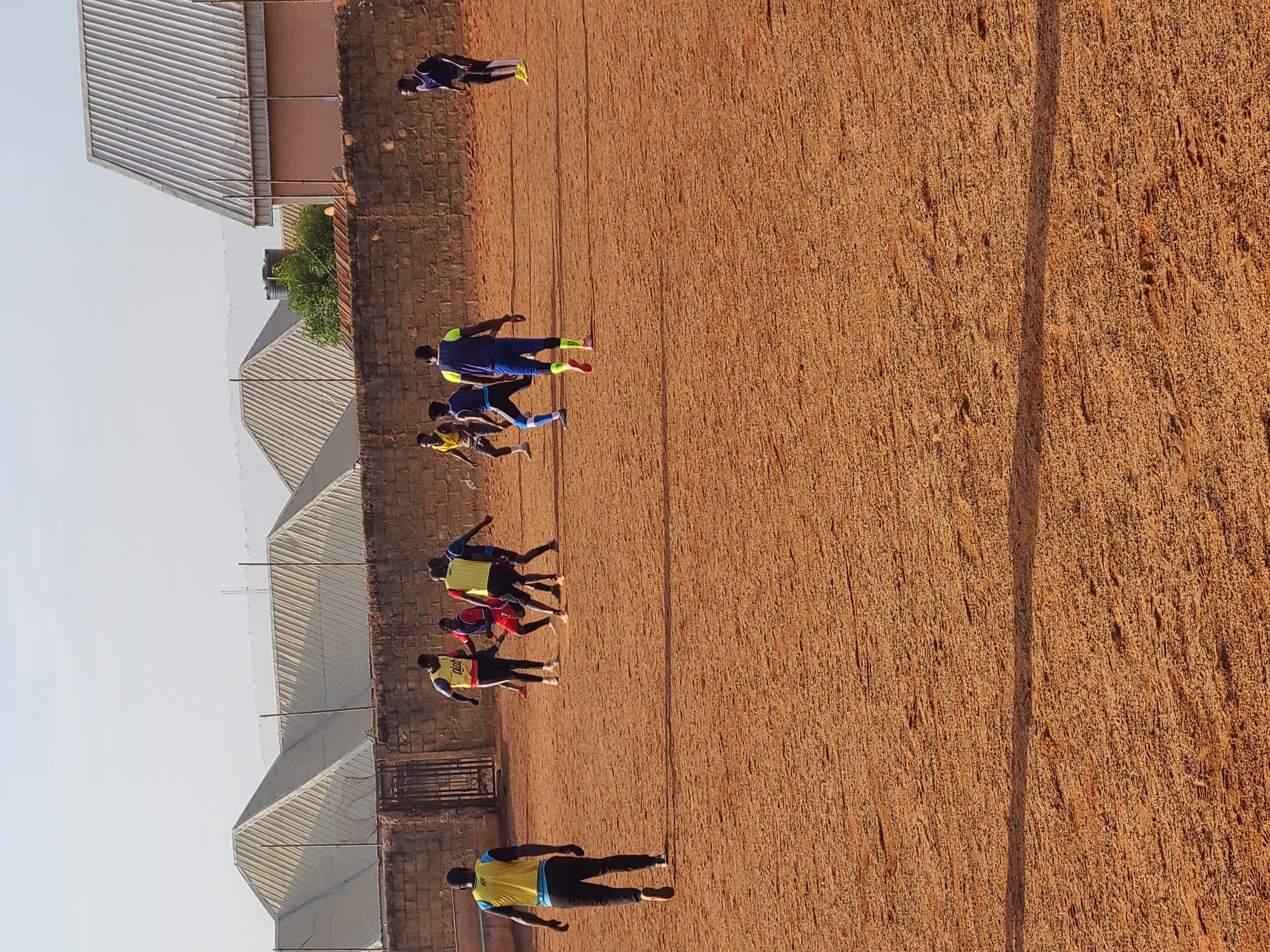 The image depicts a group of people on a reddish-brown dirt field in Barakin Lerawi, Nigeria. The group seems to be engaged in physical activity, possibly a soccer practice or training exercise, indicated by their athletic apparel. Some are running, others are standing. Buildings with corrugated metal roofs are visible in the background along with a brick wall. The image appears to be taken during daylight hours with ample sunlight.