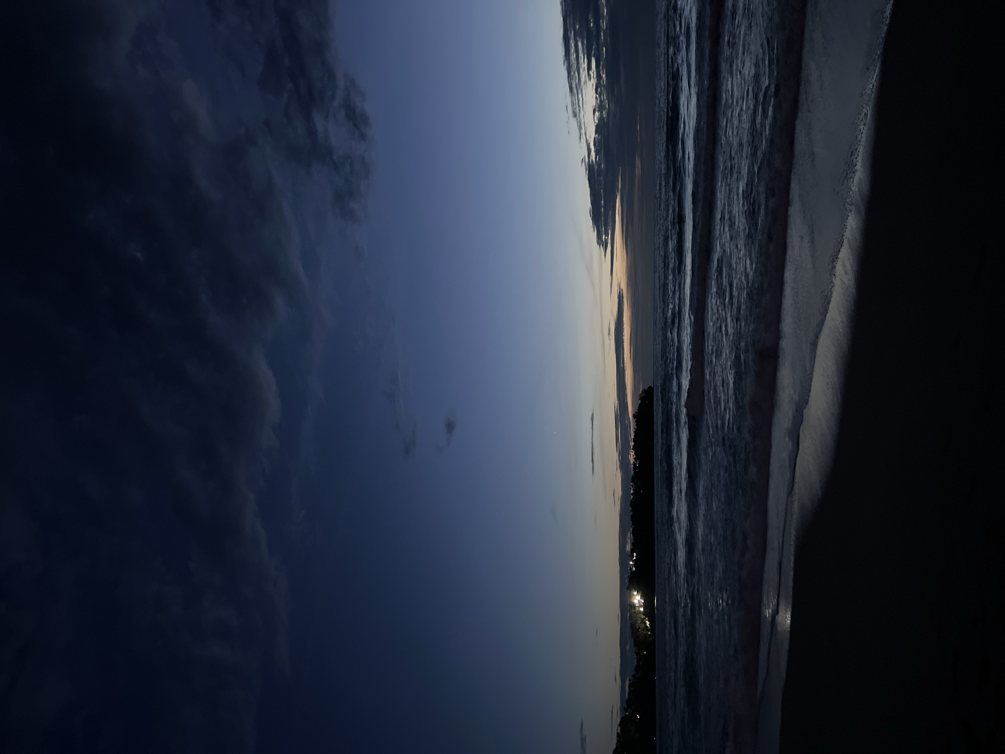 A dark, serene beach scene unfolds under a twilight sky. The foreground is dominated by the dark sand of the beach, with a faint reflection of light on the wet sand where the waves have receded. Gentle waves are breaking on the shore, their crests illuminated by an unseen light source. In the mid-ground, a dark, tree-lined landmass juts out into the ocean, dotted with several bright lights, suggesting a settlement or resort. The ocean stretches to the horizon, where it meets a sky transitioning from deep blue to a lighter hue near the horizon. Dark, wispy clouds are scattered across the sky, adding depth and texture to the scene. The overall atmosphere is one of peace and tranquility, captured during the transition from day to night.