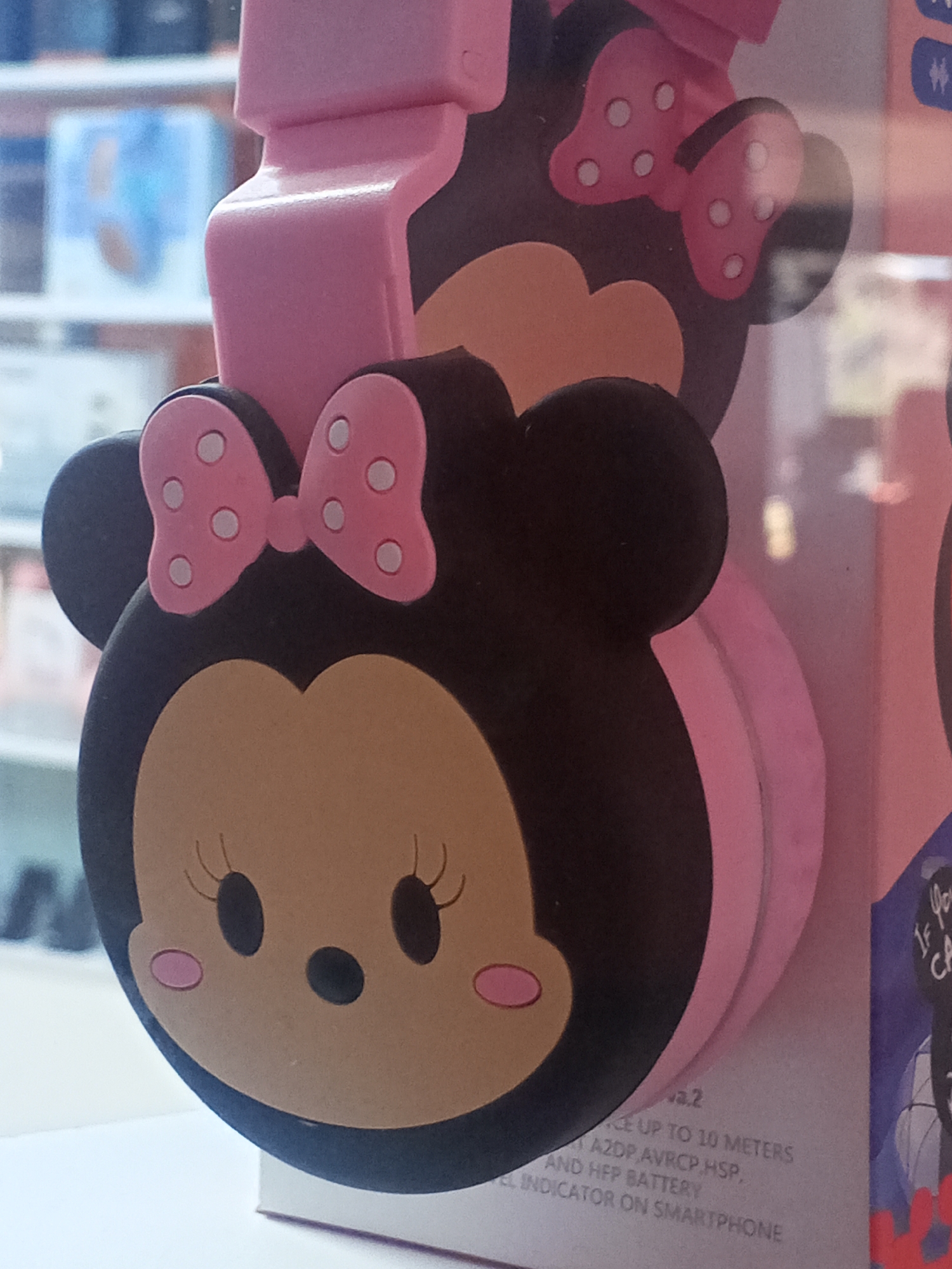 The image shows a close-up of pink headphones designed to resemble a cartoon character, possibly Minnie Mouse, with large black ears and a pink bow with white polka dots. The character's face is depicted with large black eyes, pink blush marks, and a small black nose. The headphones are a bright pink color and appear to be made of plastic. In the background, there is a blurry display of merchandise, suggesting the photo was taken in a retail environment. The location is identified as Juan Griego, Venezuela.