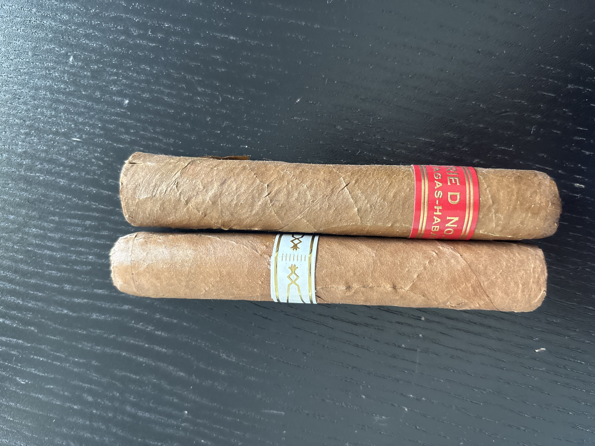 Two cigars are placed vertically on a dark, textured surface. The cigar on the left has a white and gold band. The cigar on the right has a red and gold band with text that appears to be "RED NO GAS-HAB".