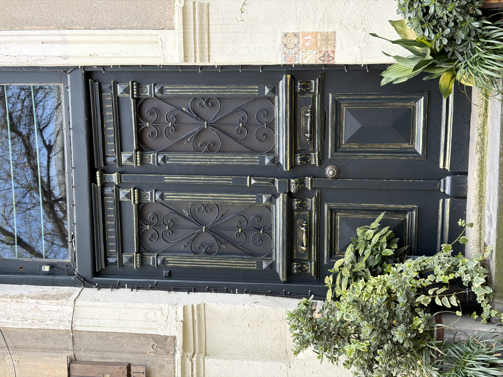 A close-up, slightly tilted, medium shot depicts an ornate, two-paneled, dark gray door with gold accents, positioned adjacent to a window on the left. The door features decorative metalwork with heart-shaped motifs on the left side of each panel, and a handle and geometric panels on the right. The door is embedded in a light-colored, textured wall, possibly stone, with visible architectural details. A small section of patterned tiles is visible above the door. To the right of the door, overflowing pots of greenery add a natural element to the scene. The reflection of bare trees is visible in the window to the left. Small white lights, possibly fairy lights, are strung along the top of the door frame. Based on the architecture, vegetation, and decorative style, the location is likely in the old city of Tbilisi, Georgia.