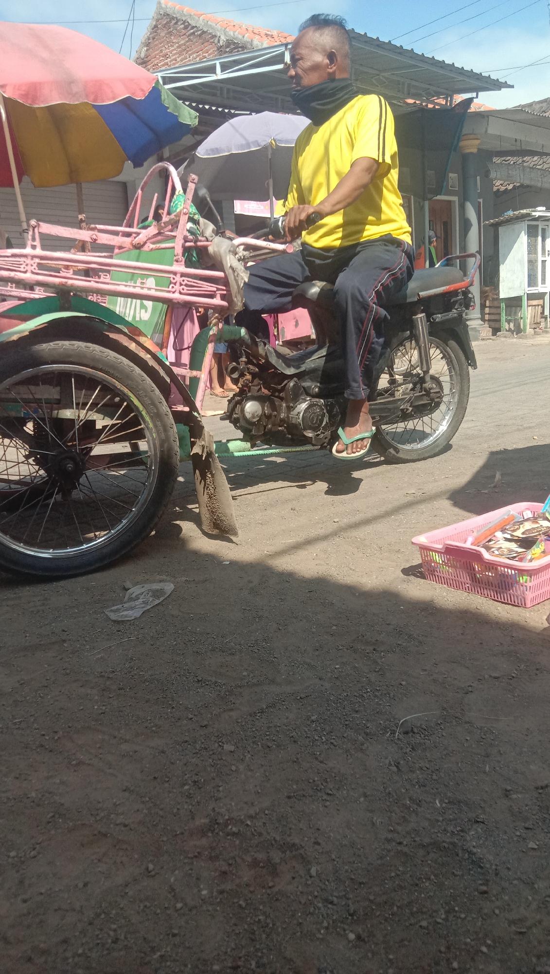 The image depicts a man riding a motorcycle with a carriage attached in Karang Anyar, Indonesia. The man, wearing a yellow t-shirt, dark pants, and a black neck covering, sits on the motorcycle, which is connected to a pink carriage. The carriage has a colorful umbrella and appears to be used for transport or carrying goods. In the background, there are buildings with tiled roofs, typical of Indonesian architecture, and a bright blue sky. The ground is a mix of dirt and gravel. To the right, a pink plastic basket filled with various items, possibly snacks or small goods, is visible. The scene appears to be a street or marketplace.