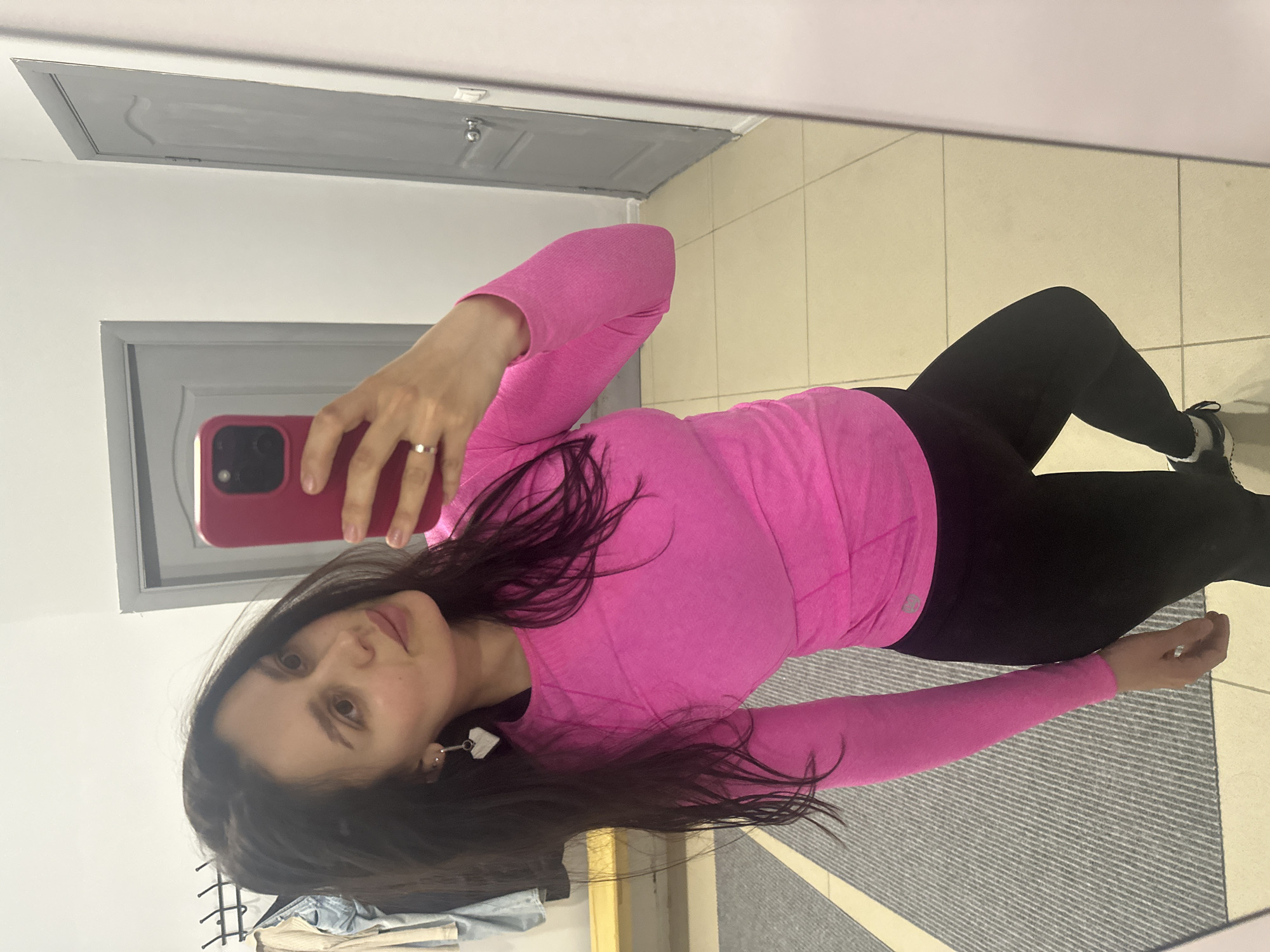 A mirror selfie taken indoors in Chaikovsky, Russia, shows a woman posing for the camera. She is wearing a long-sleeved, bright pink top and form-fitting black leggings, with dark sneakers visible on her left foot. Her long, dark hair falls over her shoulders and down her back. She is holding a red or pink-cased smartphone in her right hand, which has a silver-colored ring on her ring finger, using it to capture her reflection. A white, heart-shaped earring is visible on her left ear. She looks towards the camera with a neutral to slightly smiling expression.
The reflection in the mirror reveals a room with light-colored tiled flooring, part of which is covered by a gray, striped mat or rug. A light gray, paneled door is visible in the background, and above it, what appears to be a gray access panel or ceiling feature, suggesting a utility space or a common area. The walls are white. The overall lighting suggests an indoor setting, likely under artificial light. There is no visible text in the image.