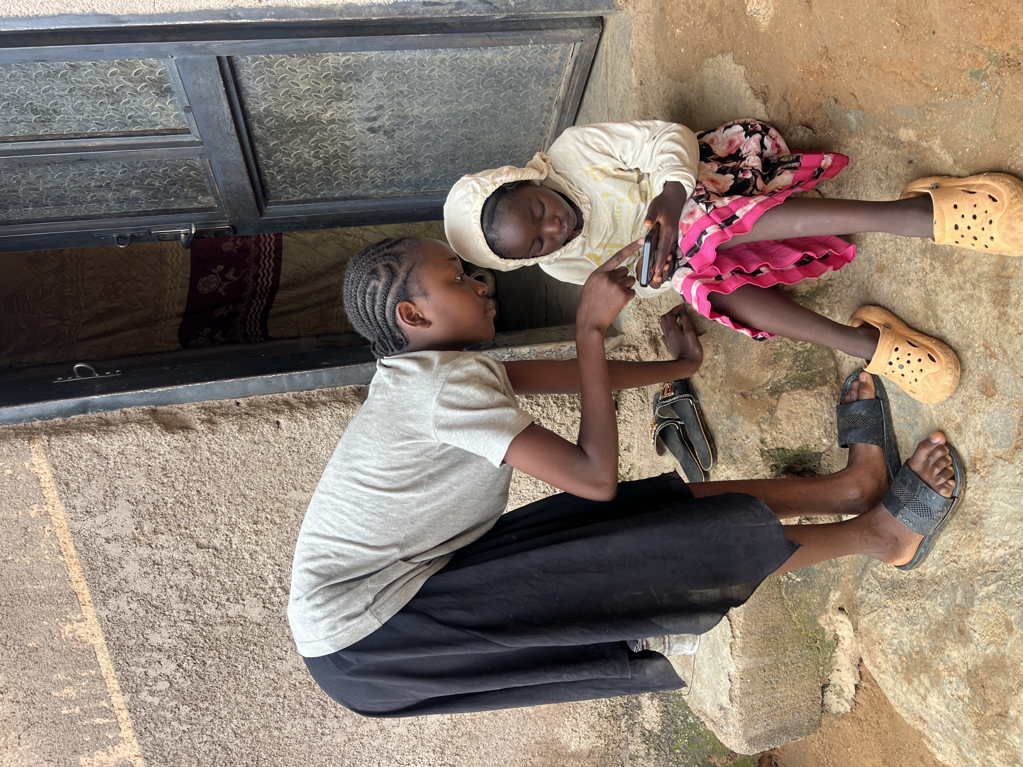 Two young Black girls are sitting on concrete steps outside a building in Jos, Nigeria. The girl on the left is standing and leaning over to point at a smartphone held by the girl on the right. The standing girl wears a grey t-shirt and a long black skirt. The seated girl wears a cream-colored hooded sweatshirt and a floral skirt with a bright pink trim. She has on light brown clog-style shoes, and the standing girl wears black slides. Behind them, a metal-framed door with glass panes is partially open, revealing patterned curtains. The building wall appears to be made of textured concrete.