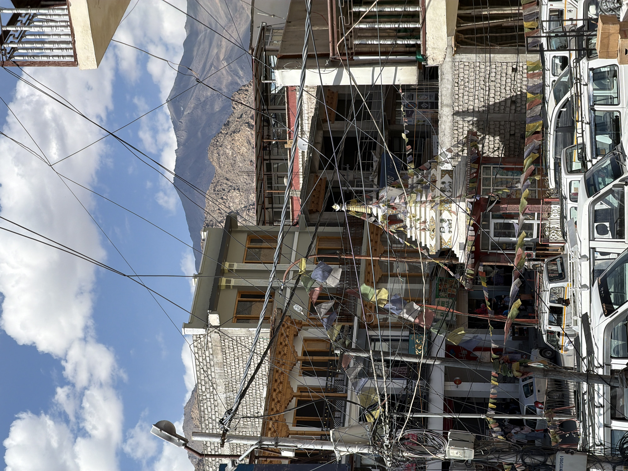 The image depicts a busy street scene in Leh, India, characterized by an abundance of overhead electrical wires crisscrossing the frame. In the foreground, several white SUVs and vans are parked, suggesting a transportation hub or a commercial area. The buildings lining the street are a mix of traditional and modern architecture, with some featuring intricate wooden carvings and others with stark concrete facades. Prayer flags, a common sight in Buddhist regions, are strung across the street, adding a splash of color to the otherwise muted tones of the buildings and landscape.
In the middle ground, a white stupa, a Buddhist religious monument, stands prominently, adorned with more prayer flags. Behind the stupa and buildings, a majestic mountain range looms in the background under a bright blue sky dotted with white clouds, indicating a clear day in the high-altitude region. The overall impression is one of a vibrant, densely populated urban environment set against a dramatic natural backdrop, with the omnipresent wires symbolizing the blend of tradition and development in Leh.