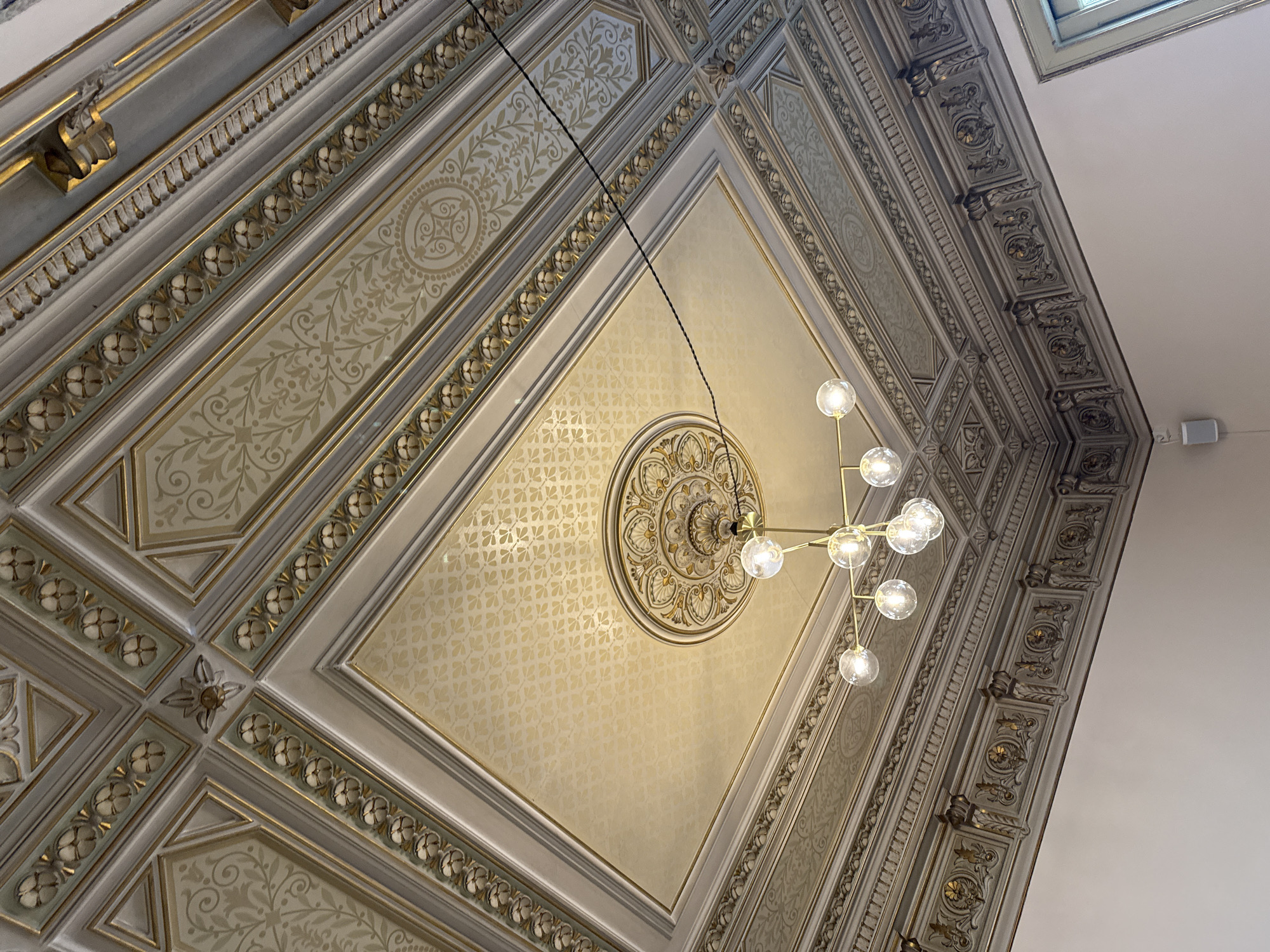 An upward-angled view captures an ornate ceiling interior. The architectural design showcases intricate moldings with decorative floral patterns, and a central ceiling medallion. A modern chandelier hangs from the ceiling's center. The location is an interior in Barcelona, Spain.