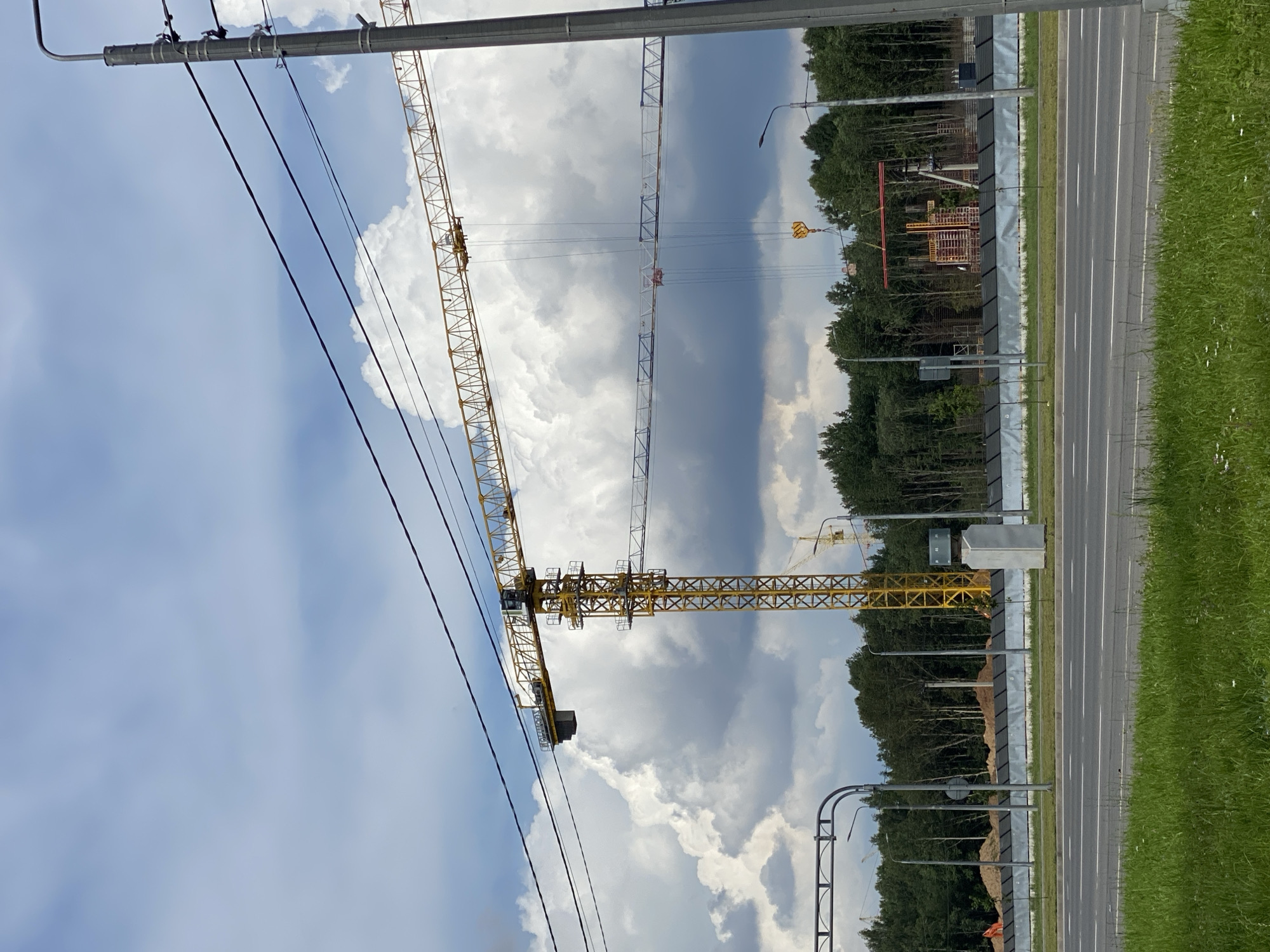 The image shows a construction site with multiple yellow and white tower cranes against a cloudy sky. In the foreground, there is a road with a grassy median and a sidewalk. The background features a dense line of trees, a construction fence, and piles of dirt. A prominent yellow tower crane is positioned centrally, with its jib extending horizontally. A second, lighter colored crane is visible behind it. In the distance, more construction activity, including what appears to be rebar cages, is present. The overall scene depicts ongoing construction in an area with natural greenery bordering the development. The specific location is stated to be the city of Cherepovets, Russia.