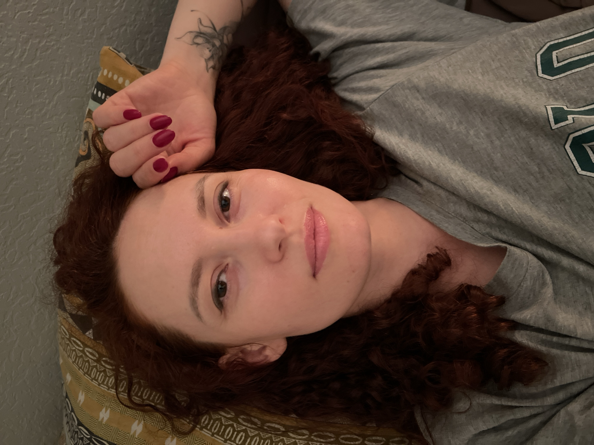 A fair-skinned woman with curly red hair is lying down, facing the camera. Her head rests on a patterned pillow. She is wearing a gray t-shirt with green lettering. Her left arm is raised above her head, revealing a tattoo on her inner arm. Her fingernails are painted dark red. The background is a light gray wall. There is no indication of this image being located in Saint Petersburg, Russia.