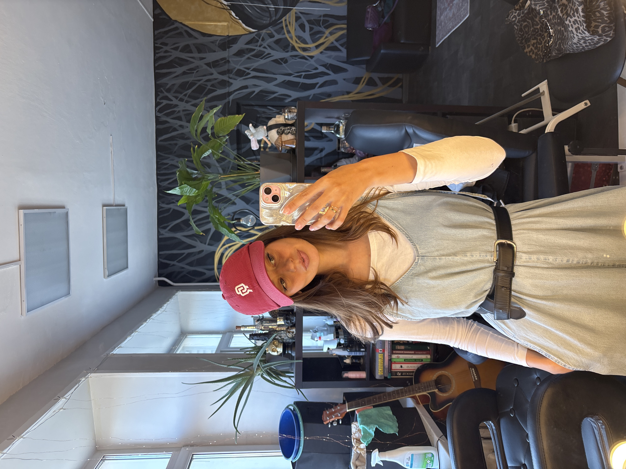 A young woman with long brown hair is taking a selfie indoors. She is wearing a red baseball cap with a white logo, a long-sleeved white shirt, and a light-blue dress with a black belt. She is holding a gold-colored phone. Behind her, there is a bookcase with books, bottles, and other objects. A guitar leans against the bookcase. A large plant is also visible. There are chairs and a mirror in the room. The location is possibly Krasnoyarsk, Russia.