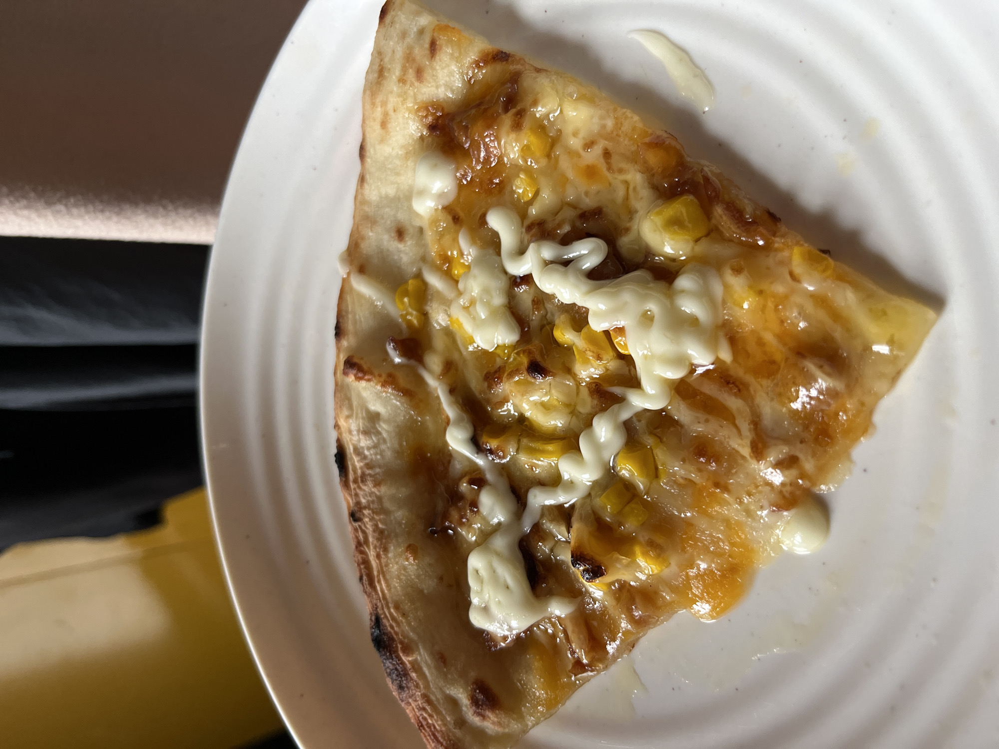A slice of corn and cheese pizza is presented on a white, ribbed plate. The pizza features a golden crust, melted cheese, and visible corn kernels. White sauce, possibly mayonnaise or a cheese sauce, is drizzled over the top in a decorative pattern. The background is out of focus, showing a yellow surface on the left and darker fabric on the right, suggesting an indoor setting. The scene is not set in a city.