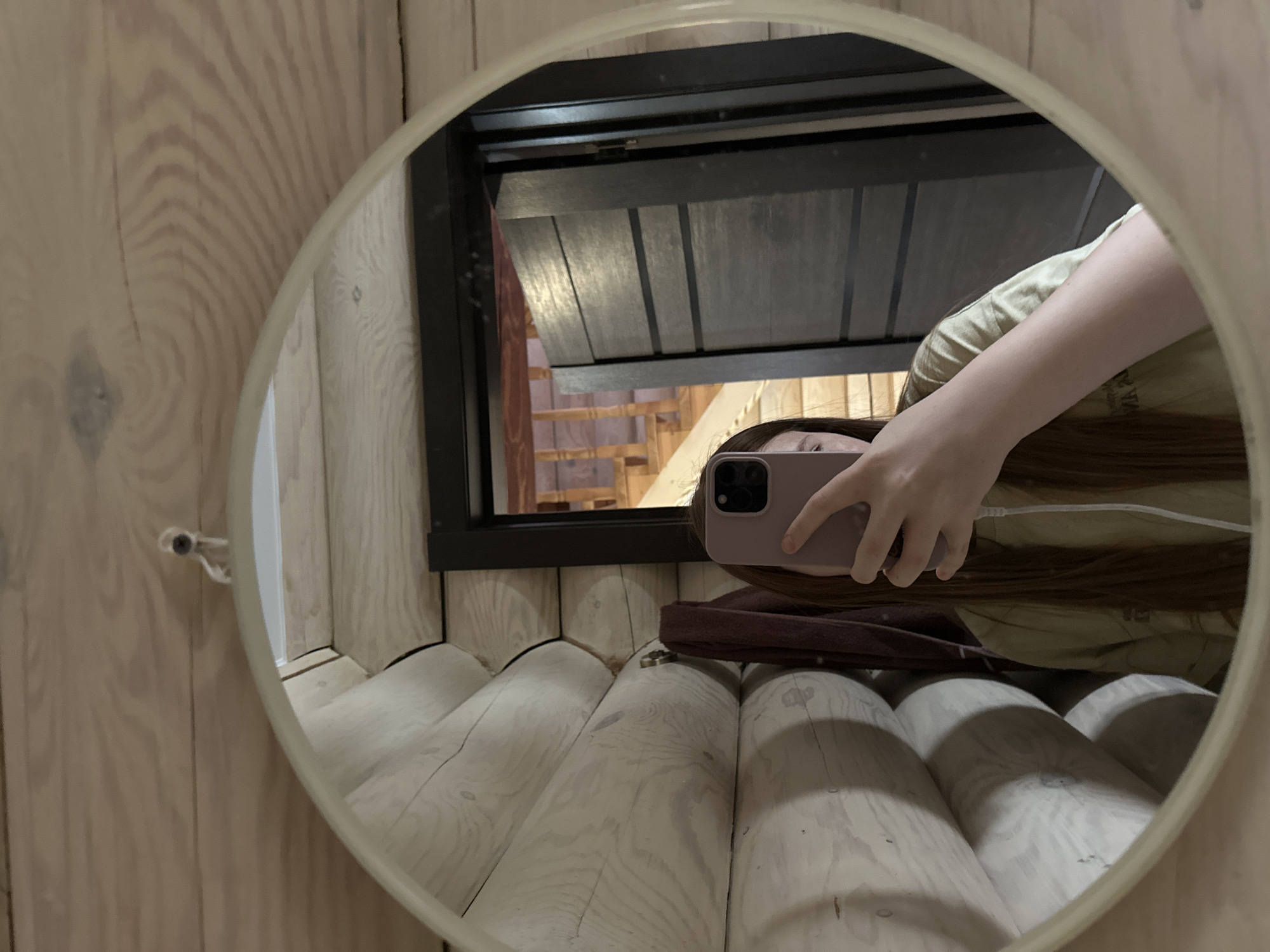 A reflection in an oval mirror shows a woman taking a selfie in what appears to be a rustic wooden cabin, possibly located in or near Ольховка, Russia. The reflection reveals part of the woman's face, her arm holding a pink smartphone, and her brown hair. She is wearing a light-colored shirt. Behind her is a small window with a partially open shutter, revealing a view of wooden stairs and more wood paneling. The wall and floor in the mirror's reflection are made of light-colored, rounded logs. The mirror itself is attached to a wood paneled wall.