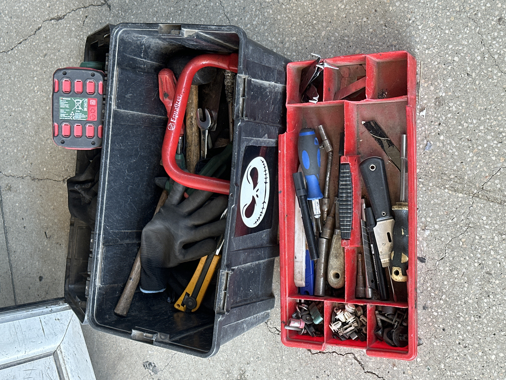 The image shows two open toolboxes on a cracked concrete surface, possibly outdoors. The larger, black toolbox on top contains a pair of black work gloves, a yellow utility knife, and a red power tool battery pack with multiple buttons. A red U-shaped tool with "Equalizer" printed on it is also visible. A graphic of Jack Skellington's face is affixed to the side of the black toolbox. The smaller, red toolbox below is filled with various tools and fasteners. These include screwdrivers with blue and black handles, various small metal parts like screws and bolts, and at least two utility knives. The scene is well-lit, suggesting it is daytime.