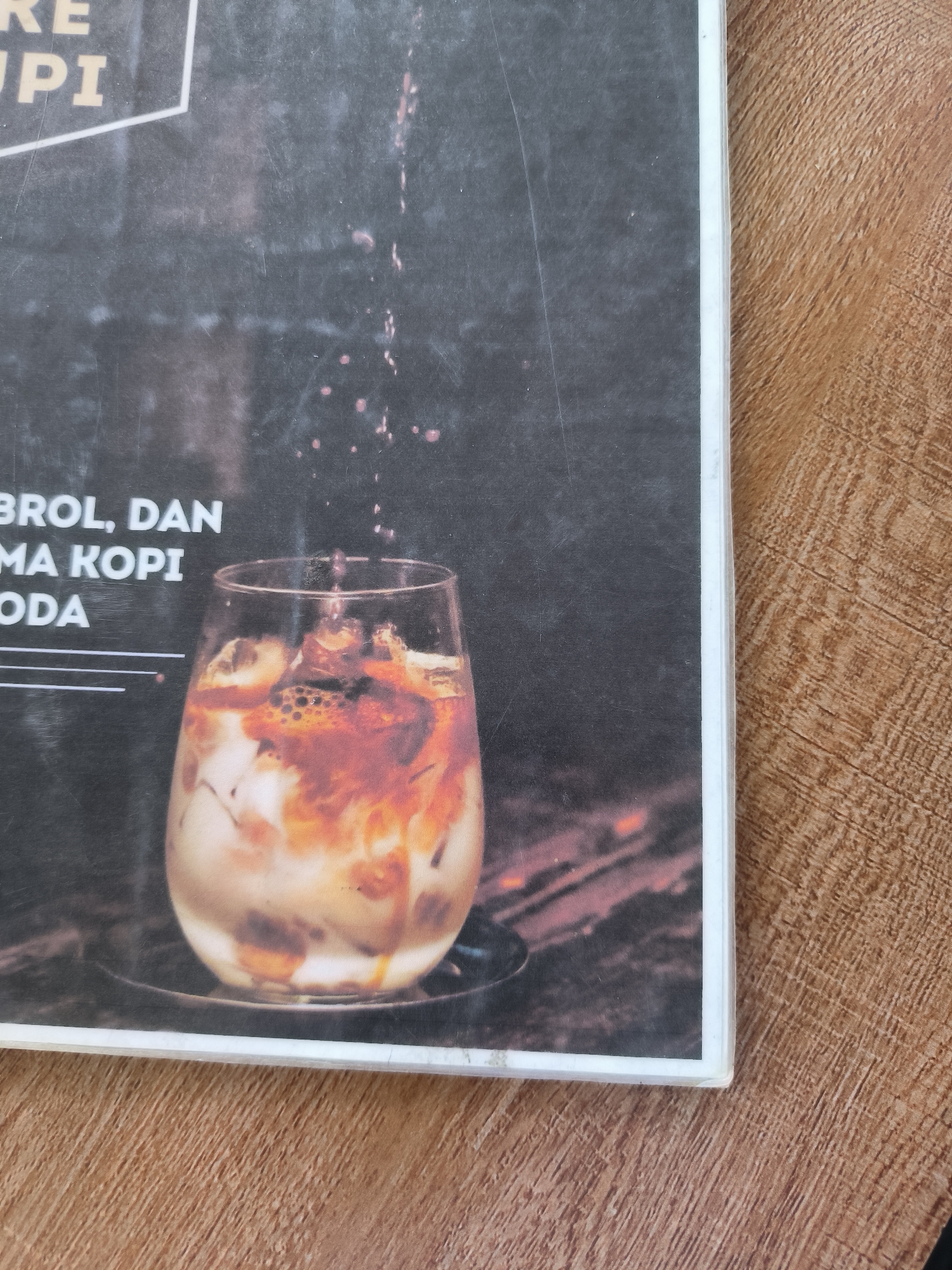 The image shows a promotional poster or advertisement placed on a brown woodgrain surface. The poster is printed with a background of dark grey to black, featuring a stylized depiction of a glass of iced coffee. The coffee beverage appears to be mixed with ice, milk, and coffee, creating a swirling effect of brown and white colors inside the glass. Small drops of a liquid are displayed above the glass, seemingly in the process of being added to the drink. The text on the poster includes partial words such as "RE UPI," "BROL, DAN MA KOPI," and "ODA," suggesting that the poster advertises a specific type of coffee or a coffee establishment. The image is likely related to the Indonesian city of Matang, and it is used to advertise a local coffee shop or beverage offering.