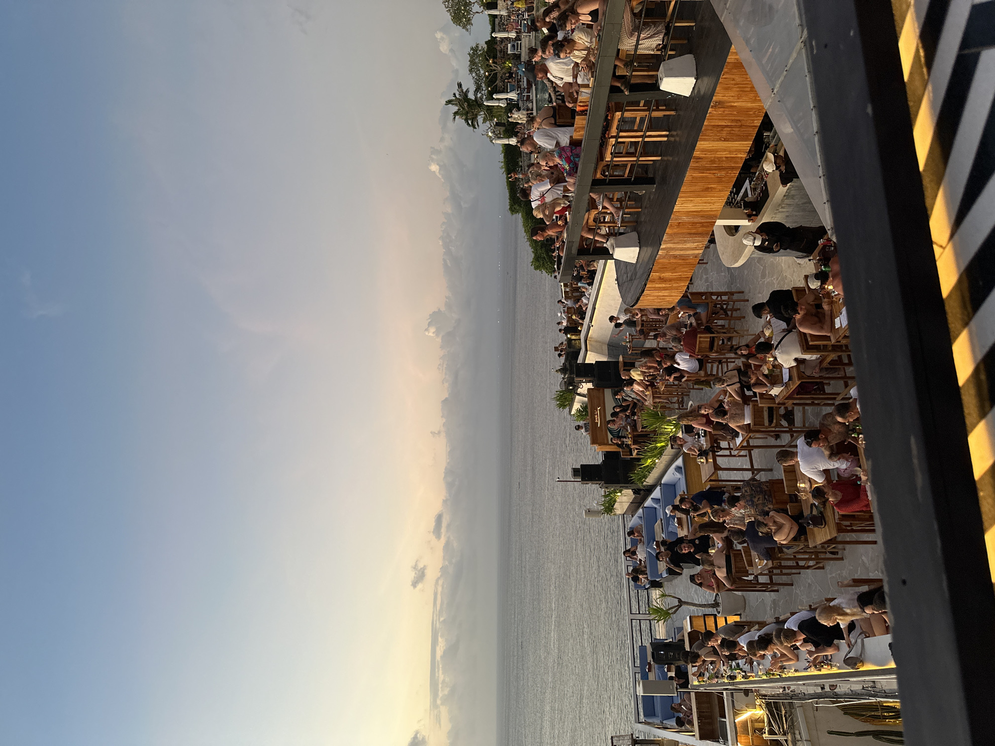 The image depicts a crowded, multi-level outdoor bar or restaurant overlooking the ocean. It is located in Kuta Selatan, Indonesia. Numerous patrons, mostly adults, are seated at tables and bar counters. They are engaged in eating, drinking, and socializing. The bar/restaurant structure includes wood decking, metal railings, and striped supports. The ocean horizon and sky are visible. The sky transitions from blue to yellowish hues, indicating sunset or twilight. Vegetation and palm trees are visible in the background. Loudspeakers and other equipment suggest live music or an event.