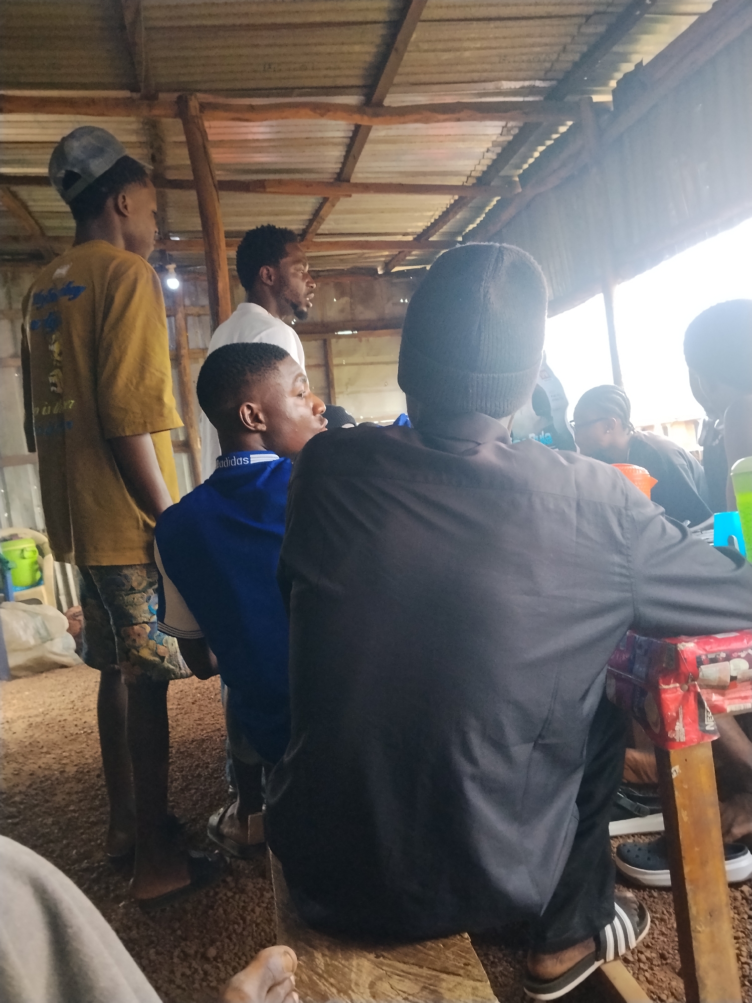 In Durumi, Nigeria, a group of young men are gathered under a rustic, open-air shelter with a corrugated metal roof supported by wooden beams. The ground is covered in gravel. One man in the foreground, wearing a black beanie and a black button-down shirt, is seen from behind, seated at a makeshift table made of wood and what appears to be recycled materials. Next to him, a young man in a blue Adidas shirt looks to his right. To the left, another young man wearing a yellow t-shirt and a grey baseball cap stands, looking in the same direction as the man in the blue shirt. Other individuals are partially visible in the background, engaged in conversation or activity. The scene appears to be a casual social gathering or meeting.