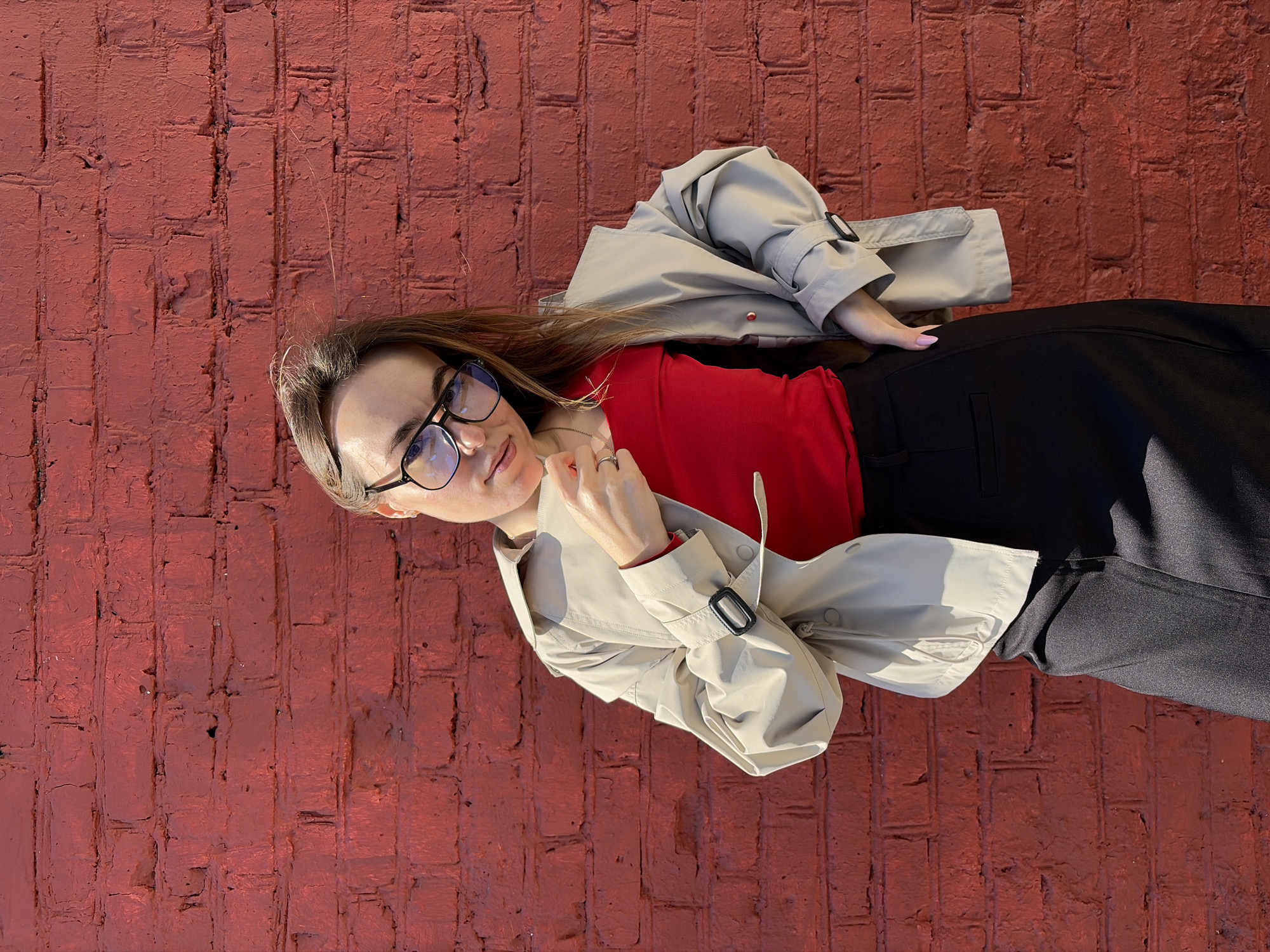 A young woman is lying on a red brick surface, seemingly outdoors. She is wearing glasses, a red top, a black skirt or pants, and a light-colored trench coat. The photo is taken from a slightly elevated angle, and the woman is looking towards the camera. The brick surface fills much of the background. The image location is unspecified, but it is not necessarily in Izhevsk, Russia. The image does not provide sufficient detail to determine if it is in Izhevsk or not.