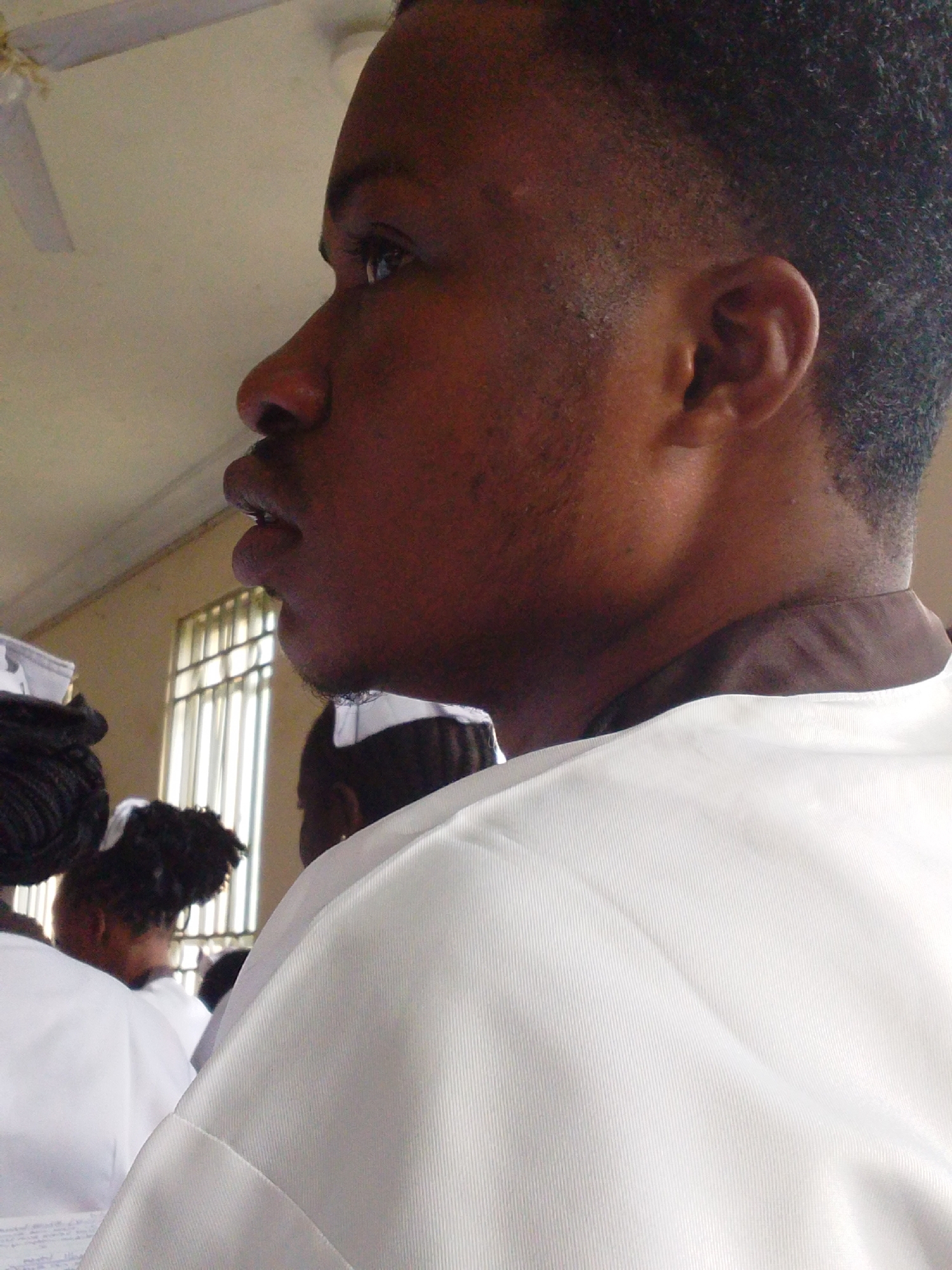The media file shows a close-up profile view of a young Black person wearing a white garment with a brown collar. The subject is looking to the left, with their mouth slightly open. Their hair is short and dark, and they have some stubble on their chin and jawline.
In the background, several other people are visible, also wearing white garments. Some have their hair styled in braids or buns. A window with vertical bars is visible in the mid-ground, suggesting an indoor setting. The lighting appears to be natural, possibly from the window.
The scene suggests a gathering or event where participants are dressed in similar attire. Given the location context of Kuson, Nigeria, it could be a school event, a religious ceremony, or a formal gathering. No specific emotions are discernible from the subject's profile, though the overall atmosphere seems calm and orderly. The image quality is moderate, with some graininess. No visible text is present in the media file.