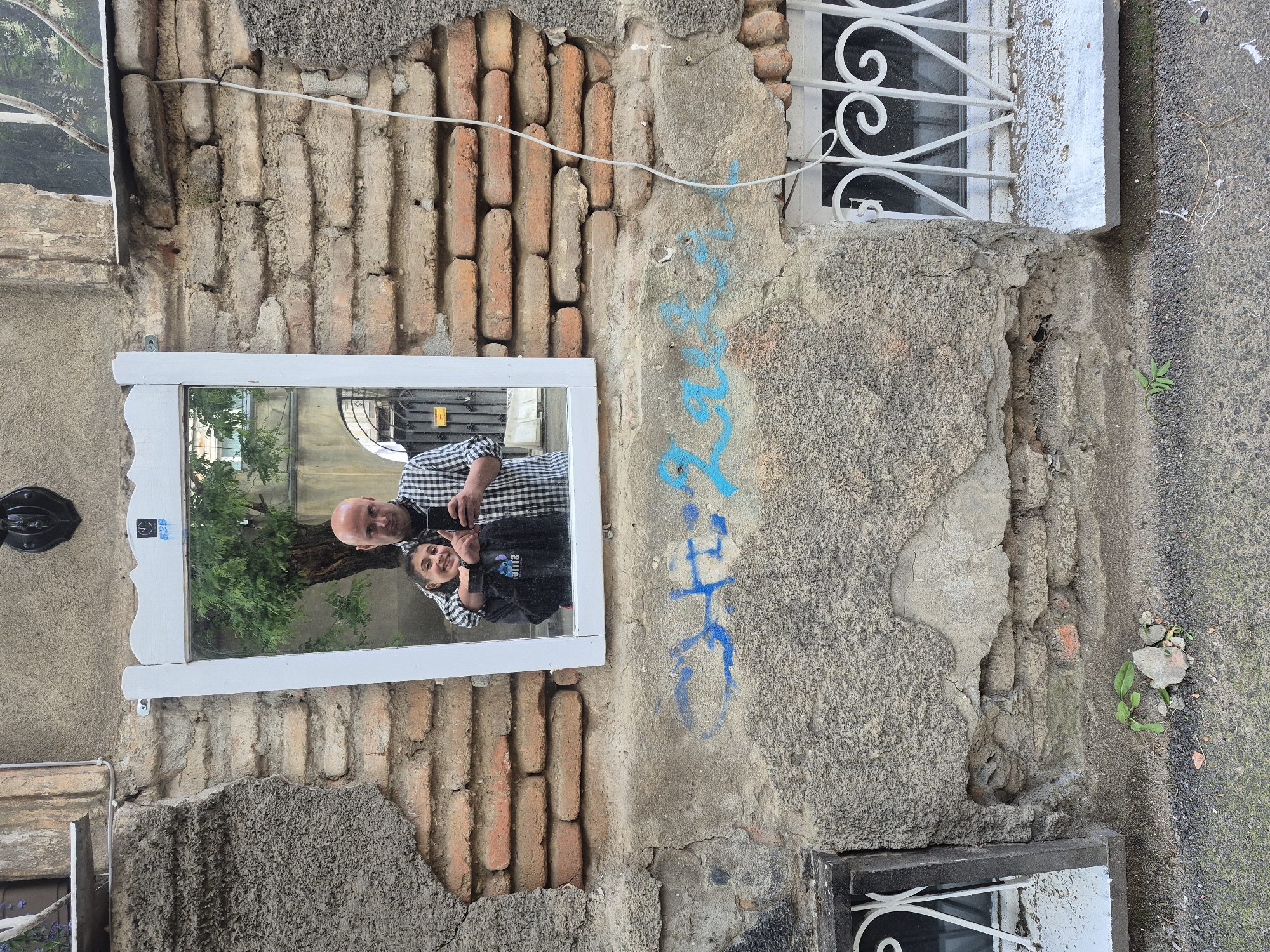 A photograph shows a section of an exterior wall, possibly in Tbilisi, Georgia, evidenced by the architectural style and building materials. The wall consists of exposed brickwork, patched stucco, and weathered paint. Graffiti, a blue design, adorns a stuccoed area. A white-framed mirror is affixed to the wall, reflecting a man and a girl taking a selfie. The man is bald, wearing a checkered shirt, and holding a mobile phone. The girl is smiling, wearing a black shirt. The reflection captures a tree and a glimpse of the surrounding environment. The wall also features window frames, one partially visible at the top of the image, and another near the bottom. The wall is weathered, with signs of damage and repair. Concrete pavement is adjacent to the wall's base.