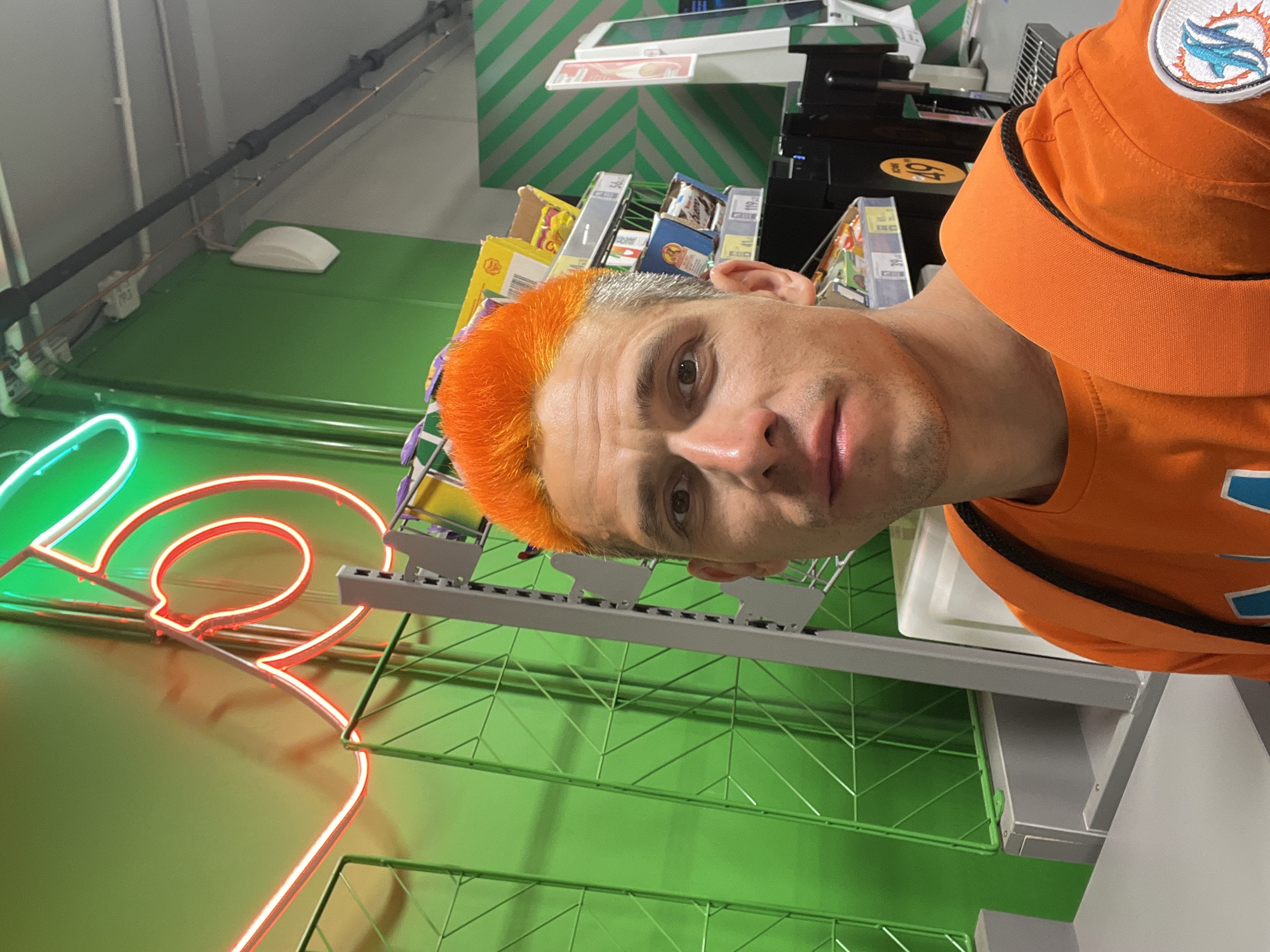 The image shows a close-up of a man with orange hair wearing an orange Miami Dolphins hat and shirt. The background is a green wall with a red and green neon sign. There is a shelf with various products on it behind the man. Based on metadata from previous queries, the location of this image is believed to be in the city of Chaikovsky, Russia.