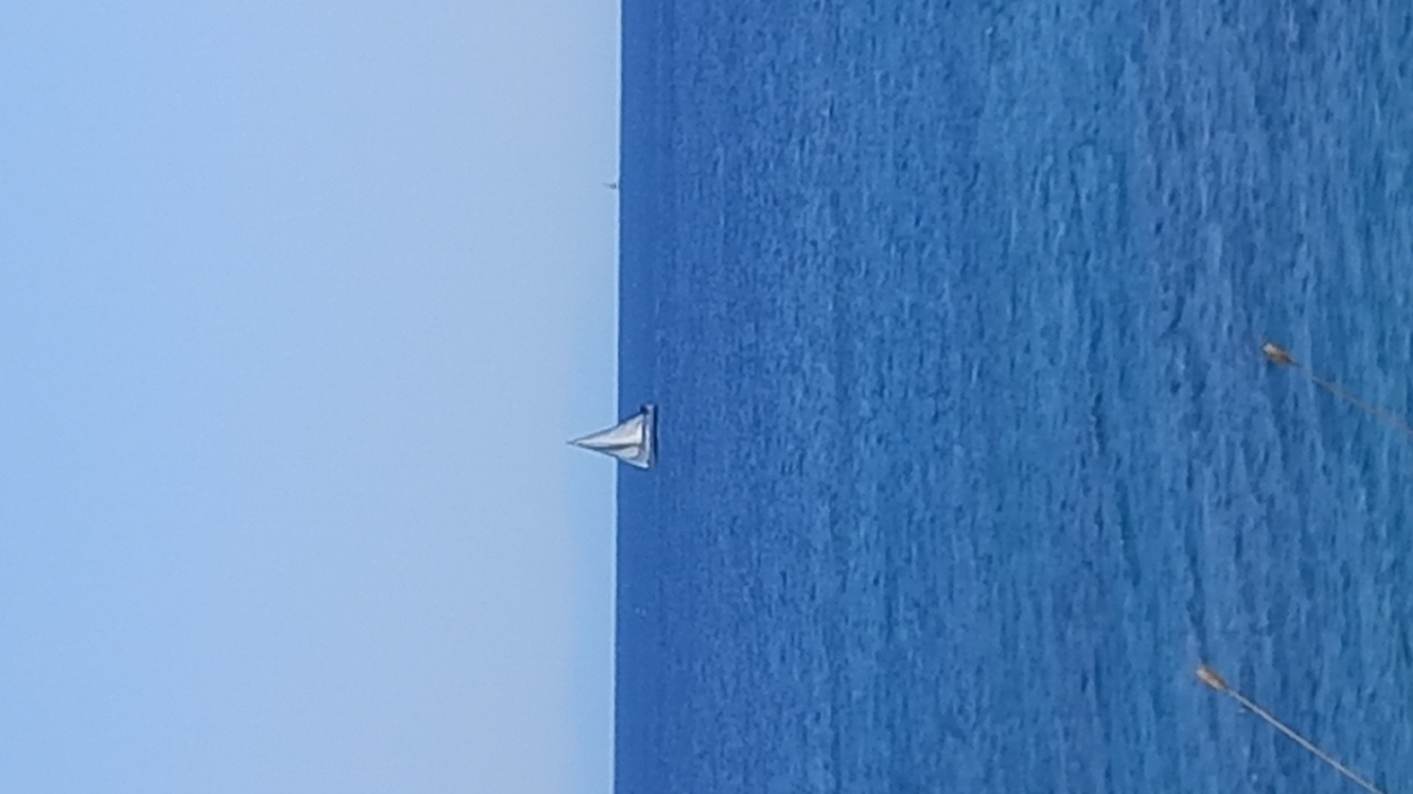 A sailboat with white sails is sailing on the Aegean Sea in the city of Kos, Greece. The water is a deep blue, and the sky is a clear, pale blue. The horizon is visible, separating the sea from the sky. In the foreground, two lampposts are visible, partially obscuring the view of the water. The sailboat is the central subject of the image, positioned slightly off-center to the left. Another smaller boat is visible in the distance on the right. The scene depicts a serene day at sea.
