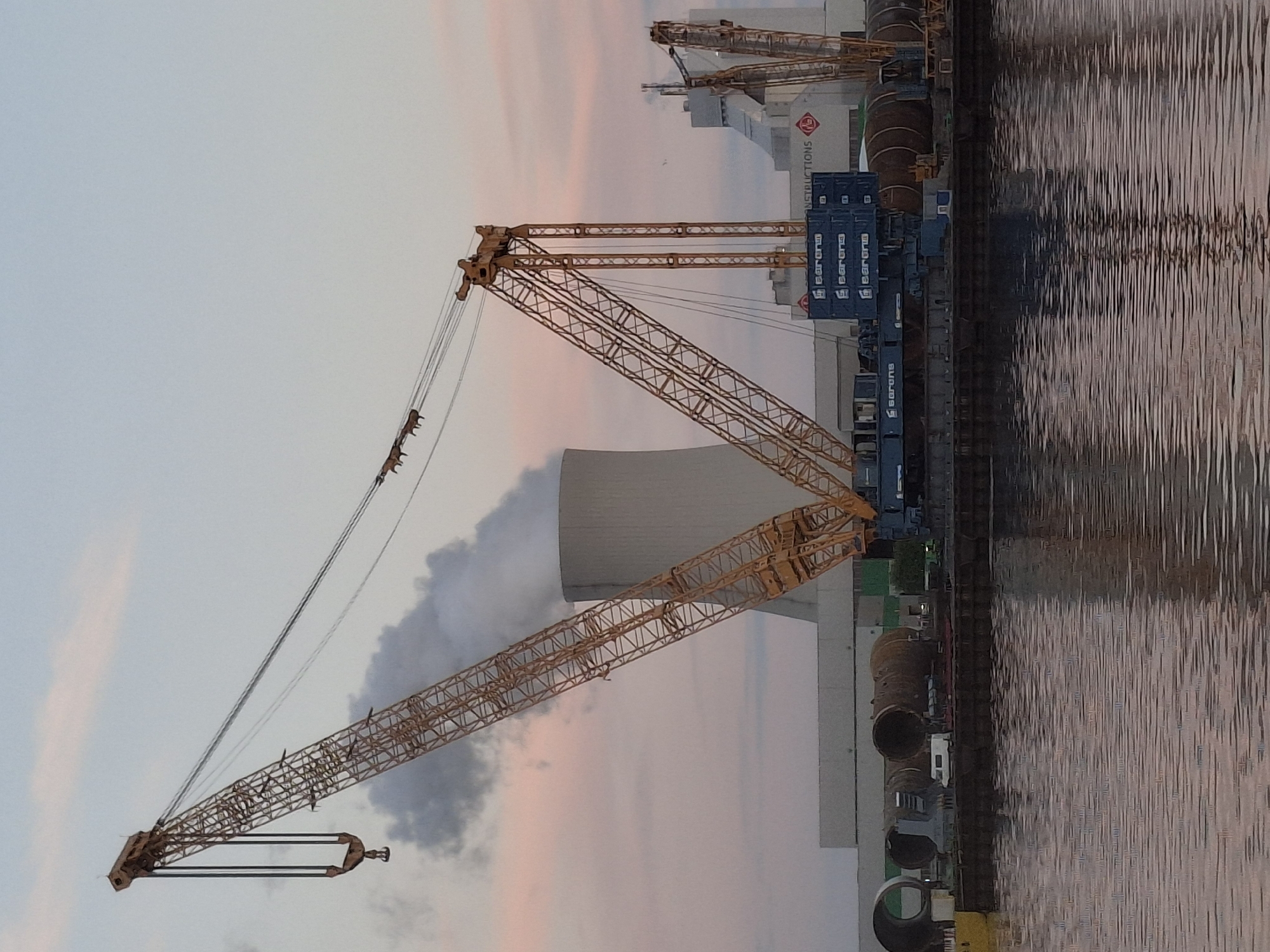The image shows a construction site on the edge of water, likely a port, in Rostock, Germany. Several large yellow cranes dominate the scene. Behind the nearest crane is a large cylindrical structure. Shipping containers stacked on the water's edge can be seen. The water reflects the sky, which is colored with shades of pink and blue at sunset.
