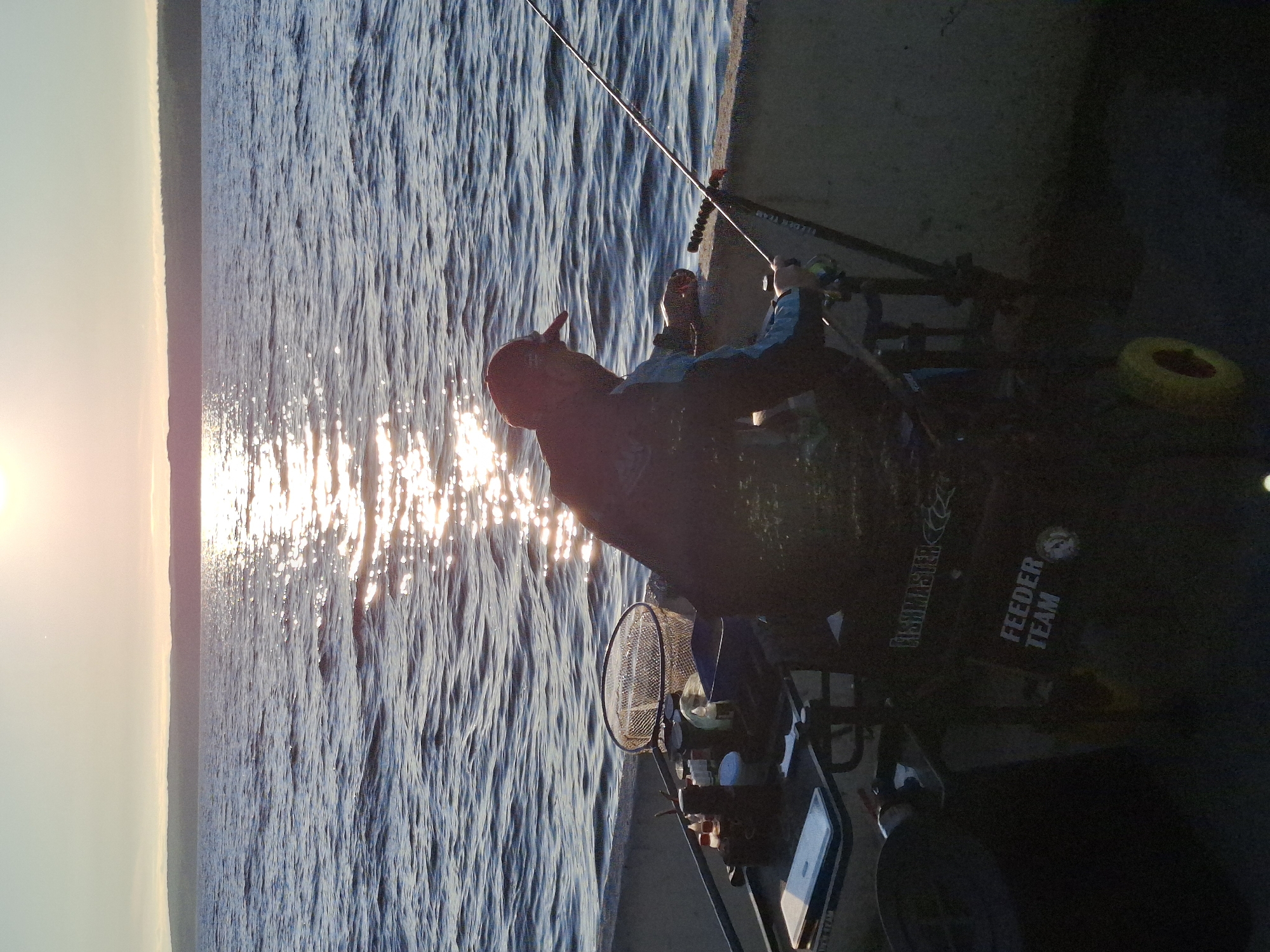 A man is fishing from a concrete embankment as the sun sets over a large body of water. The sun's reflection shimmers on the rippling surface of the water. The man, wearing a baseball cap and a fishing vest, holds a fishing rod, with his gaze directed towards the water. To his left, a fishing cart is laden with various items, including a net and containers. The scene appears to be a tranquil evening on a reservoir or large lake. It is not possible to determine the specific location within the city of Chaikovsky, Russia based on the image alone.