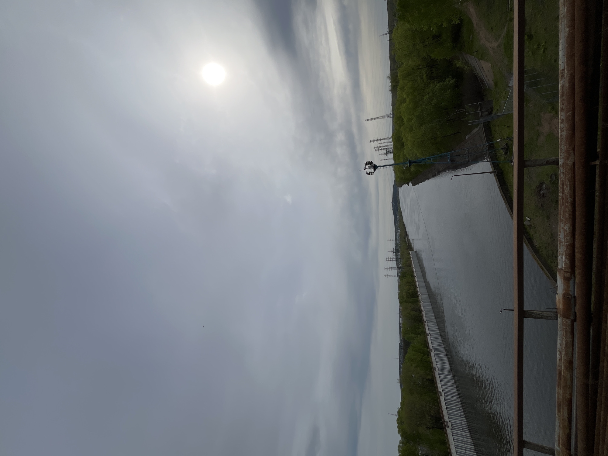The image presents a landscape scene taken from an elevated perspective, likely a bridge. A body of water, potentially a canal or river, stretches horizontally across the middle of the frame. On the right side of the water is a green landscape. A structure is on the left bank of the canal. Power lines stretch toward the horizon. The sky is overcast, with the sun faintly visible through the clouds. The composition suggests a location in an industrial or infrastructural area. The city of Chaikovsky, Russia is referenced.