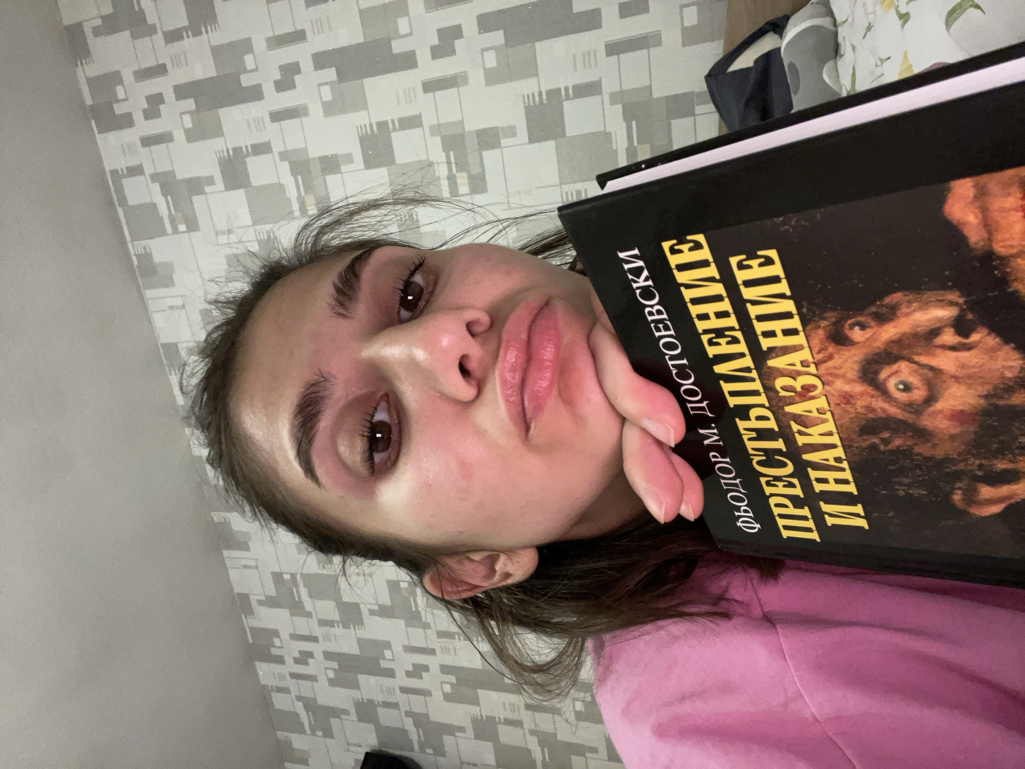 A selfie-style, tilted portrait shows a young woman holding a book close to her face. She has light skin, brown eyes, and dark brown hair. She wears a light pink top and rests her chin on her left hand. The book has a black cover with gold Cyrillic text and an image of a face. The Cyrillic text includes "ФЁДОР М. ДОСТОЕВСКИ" and "ПРЕСТУПЛЕНИЕ И НАКАЗАНИЕ". The background consists of a gray and white patterned wall. The location is not discernable from this image.