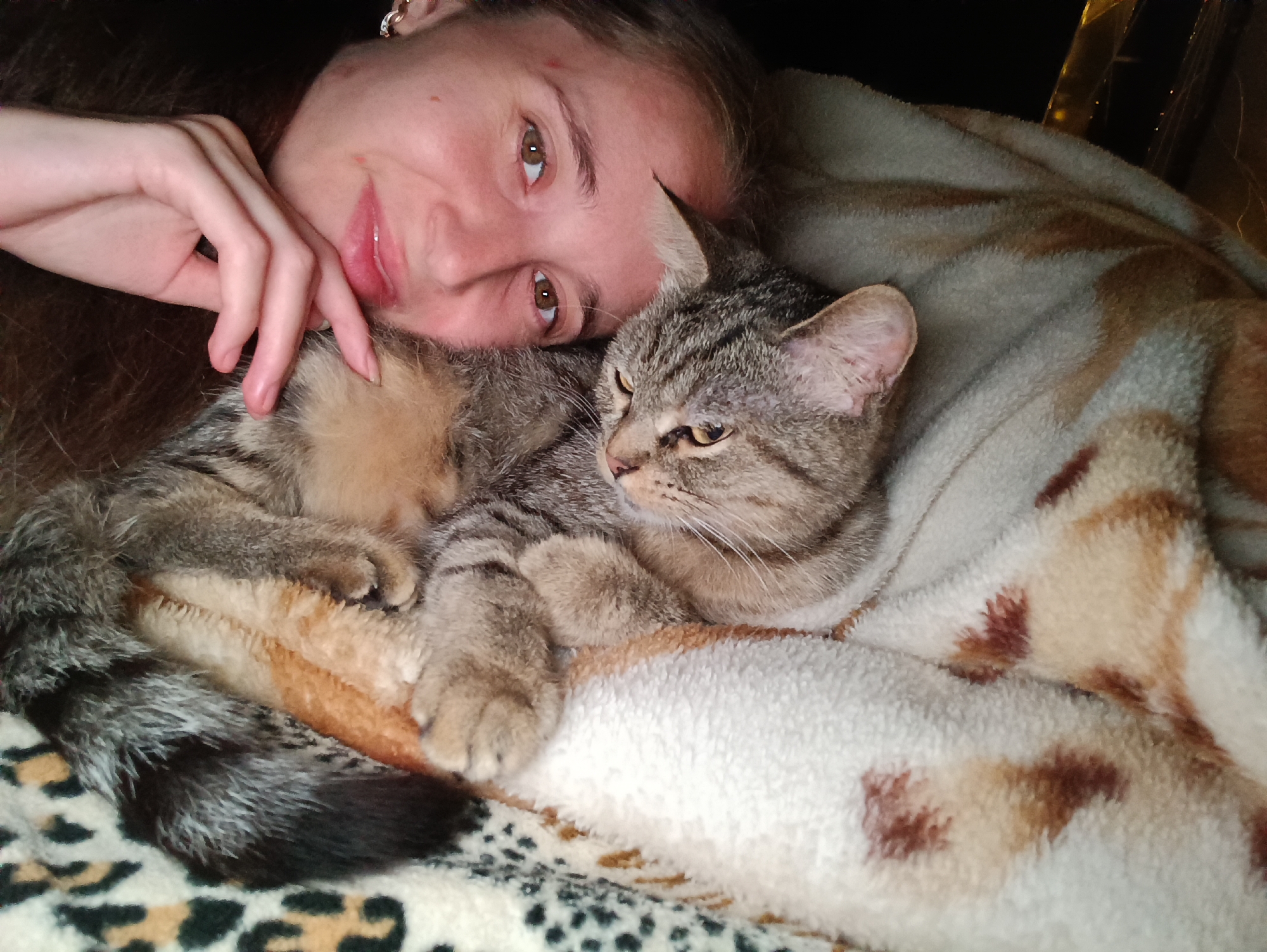 In Vypasne, Ukraine, a young woman with long brown hair and a faint smile rests her head on her arm, looking towards the camera. Her left hand is positioned near her mouth. Beside her, a tabby cat is nestled comfortably, its head resting on the woman's chest. Both are covered by a soft, patterned blanket. The background is dark, obscuring details of the surroundings.