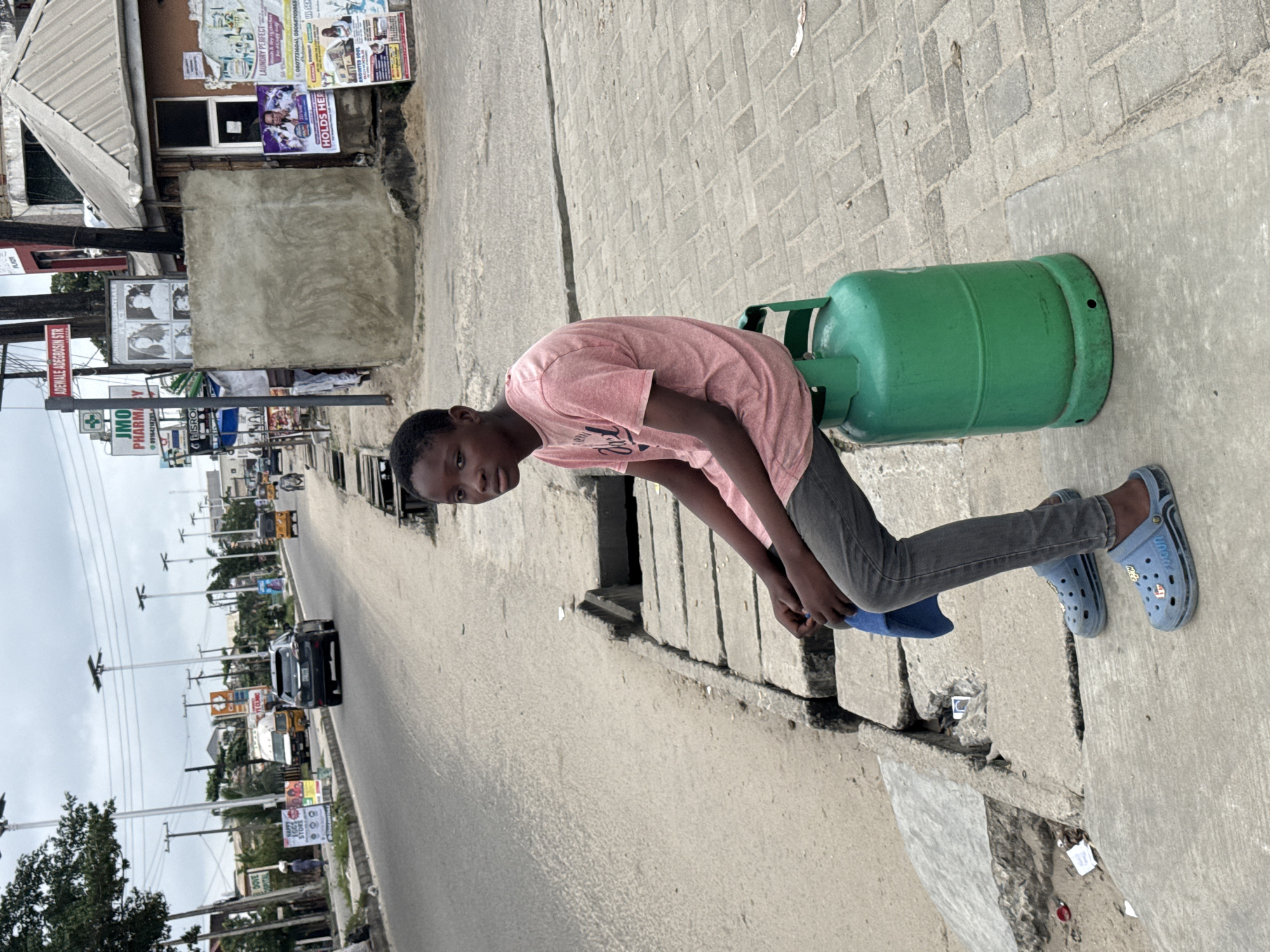 A young boy sits on a green gas cylinder on a concrete sidewalk beside a paved road in Ado, Nigeria. The boy is wearing a pink t-shirt and grey pants, with blue clogs on his feet. He holds a blue cloth in his lap. The road has several vehicles, including cars and a tricycle, and there are buildings and utility poles lining the street. Advertisements are visible on some of the buildings. The sky is overcast.