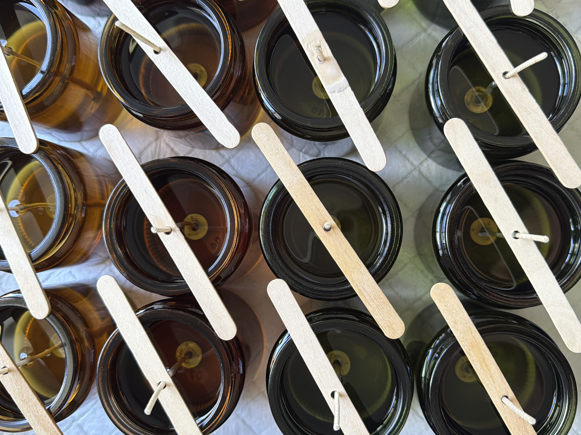 This image shows a close-up, overhead view of several amber glass jars, each containing a candle with a wooden wick holder. The jars appear to be arranged in rows on a white, textured surface. Some jars are filled with a light amber liquid, while others contain a darker, possibly green, liquid. The lighting suggests an indoor setting, possibly a workshop or production area. There are no people or any recognizable landmarks of Aespa, Estonia in the image.