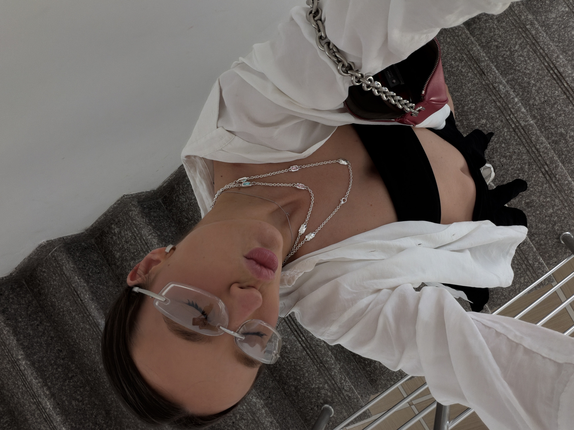 In Budva, Montenegro, a young woman with dark hair pulled back is captured from a high-angle perspective on a carpeted staircase. She is wearing clear, oversized glasses, a white unbuttoned shirt revealing a black bandeau top, and a silver necklace with colorful gemstones. A small, red handbag with a prominent silver chain strap is held in her right hand. Her lips are parted, and she is looking down, suggesting a posed selfie. The background consists of a dark gray carpeted staircase with a silver railing.