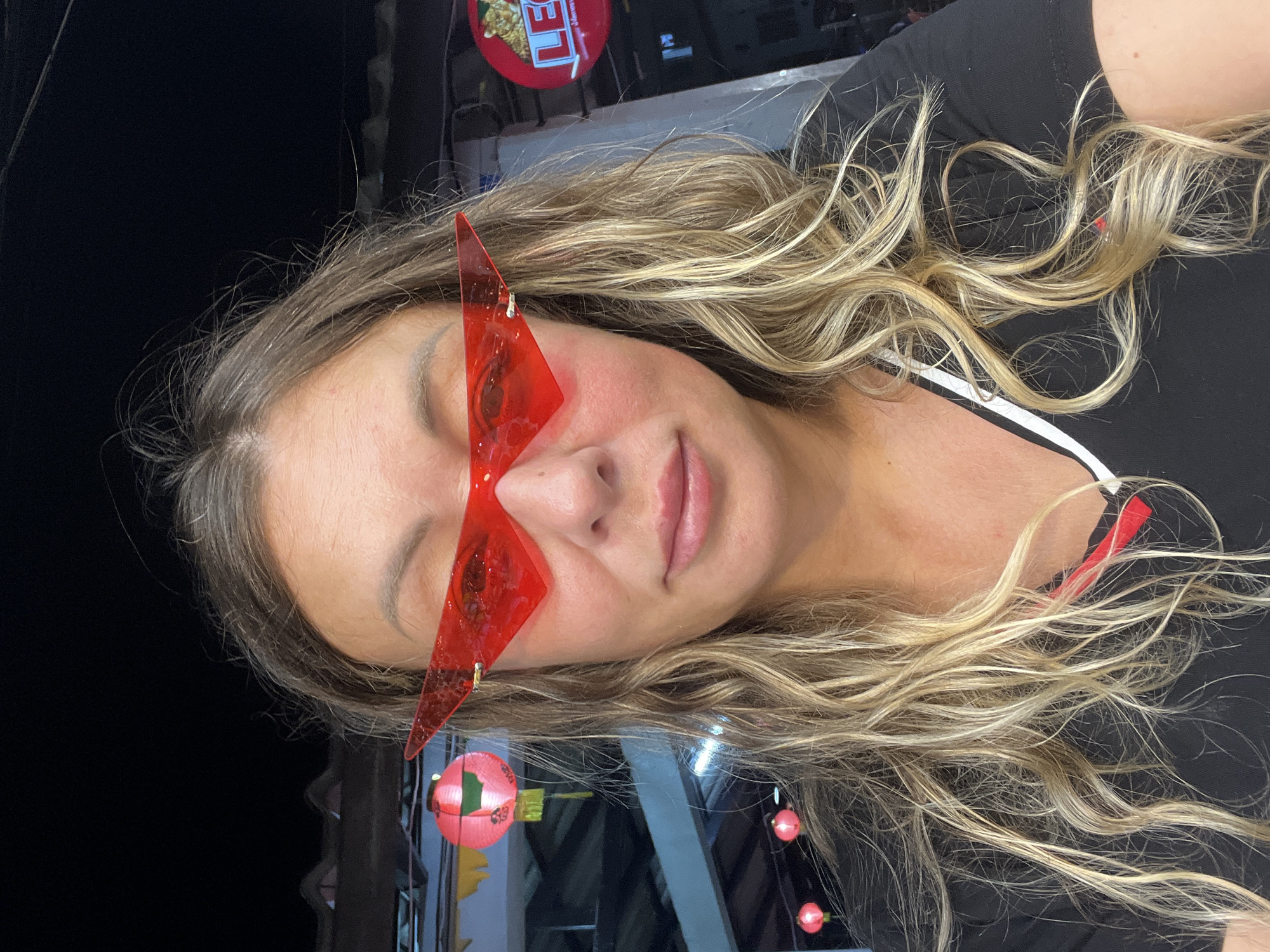 A woman with long, blonde, wavy hair is taking a selfie, wearing unique, angular, red-tinted sunglasses. She is wearing a black t-shirt with white and red trim. Her face is slightly flushed. The background appears to be outdoors at night, with a hint of a building or stall and decorative lanterns, suggesting a festive atmosphere. The overall mood is casual and playful. The location context is Ko Pha Ngan, Thailand, which is known for its full moon parties and vibrant nightlife, consistent with the scene depicted. There is some text visible on a red circular sign in the upper left of the image, but it is too blurry to read.