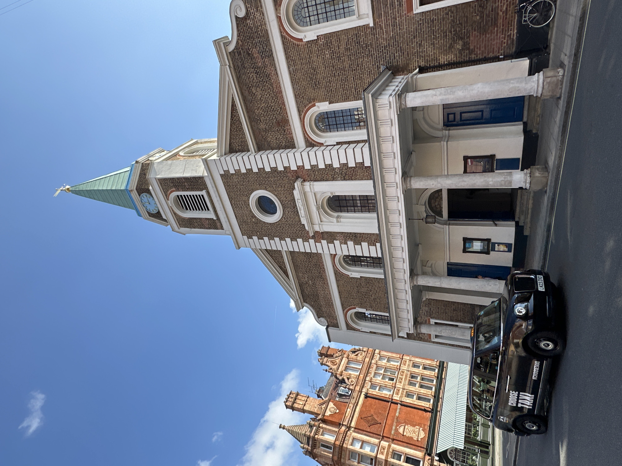The image shows a street scene in London, United Kingdom, featuring St Anne's Church, Limehouse. The church, constructed of brick with white architectural accents, is the prominent structure in the photo. A black London taxi cab is positioned in front of the church. A red brick building with ornate architectural detailing is seen in the foreground, partially obscured by the taxi. The sky is blue with scattered clouds.