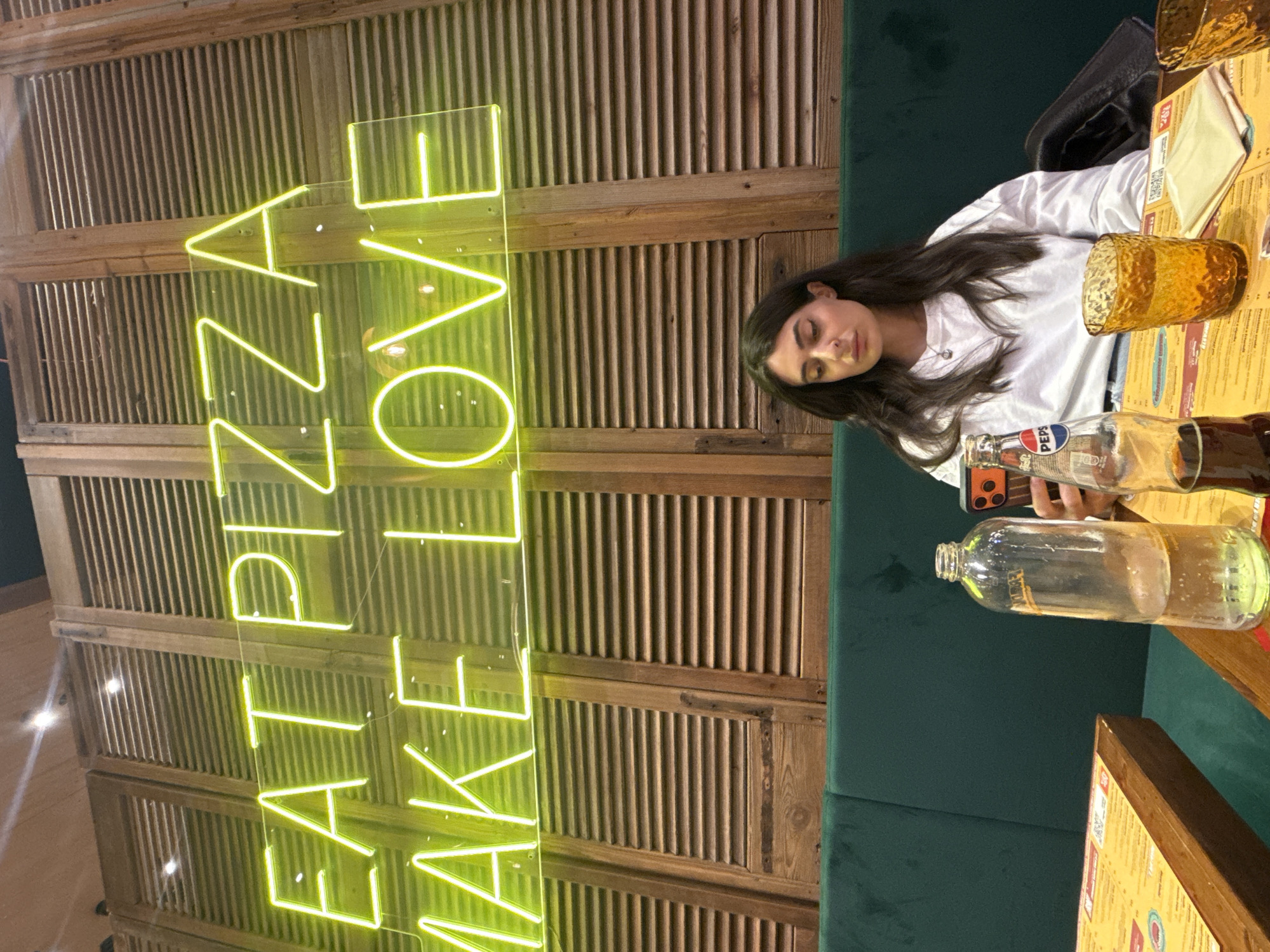 A young woman with long brown hair sits at a table in a restaurant, seemingly resting or sleeping with her eyes closed. She is wearing a white top and appears to be in a booth with green upholstery. On the table, there is a menu, a drinking glass, a bottle of Pepsi, and an empty water bottle. A neon sign that reads "EAT PIZZA MAKE LOVE" is on the wall behind her. Given the presence of an Italian brand soft drink and the sign’s message, the location is likely a restaurant in Italy. The restaurant's decor includes wooden slatted panels on the wall. Assuming the presence of Pepsi and the neon sign are reliable clues, it is most probable that the location of this restaurant is in Milan, Italy.