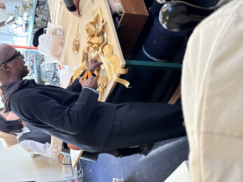 This image captures an indoor workshop or classroom scene in Arcueil, France. In the foreground, an adult male with dark skin and a shaved head, wearing glasses, a dark jacket over a dark hooded top, and dark trousers, stands at a light-colored wooden table. He is engaged in cutting a length of gold-colored ribbon with orange-handled scissors. The table surface, covered with a light brown material, is strewn with multiple cut pieces and partially formed loops of the same gold ribbon.
To the left of the male, partially obscured, is an adult female with dark hair, wearing a white collared shirt beneath a dark grey pinafore-style garment. In the background, a light-colored dress form and two industrial sewing machines are visible, indicating a textile or craft environment. A clear plastic container on the table behind the male holds several light-colored, circular objects. Rolls of dark fabric are stored beneath the table, which features green metal legs. An open wooden drawer, positioned to the right of the male, contains various spools of thread and craft supplies. A third person's hand is visible on the far right edge of the table, near a roll of gold ribbon. The floor is covered with a dark blue carpet.