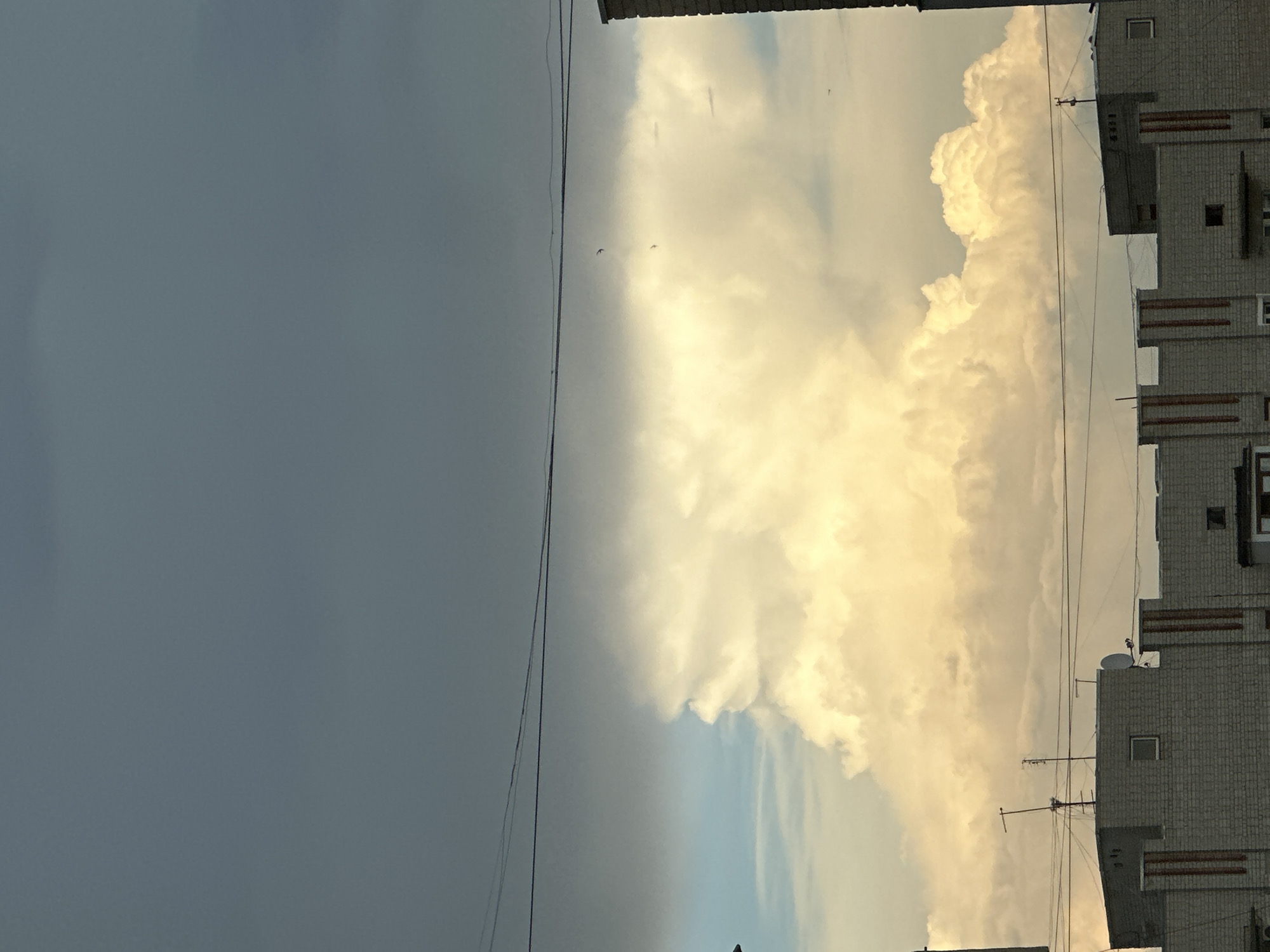 The image captures a sky scene above urban rooftops in an unknown city. Dominating the upper portion of the frame are dark, ominous storm clouds, with a sliver of lighter sky visible on the left. Below this, a massive, brightly lit cumulus cloud formation catches the light, appearing almost incandescent against the darker sky. The cloud's shape is dramatic and voluminous, suggesting turbulent atmospheric conditions.
In the foreground, the tops of several multi-story buildings are visible. These structures appear to be made of light-colored brick, with architectural details like vertical striped patterns and small rectangular windows. Various antennas and satellite dishes are mounted on the rooftops, indicating residential or office buildings. Overhead wires stretch across the sky, adding to the urban landscape. Two small birds are depicted in flight against the backdrop of the illuminated cloud. The overall impression is one of a dramatic sky event unfolding over a typical city environment.